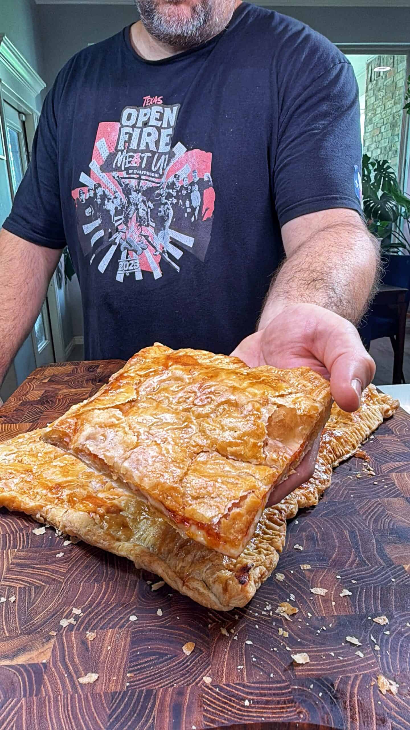 A person wearing a black T-shirt is holding a large, square piece of golden, flaky pastry over a wooden cutting board with more pastry pieces in the background.