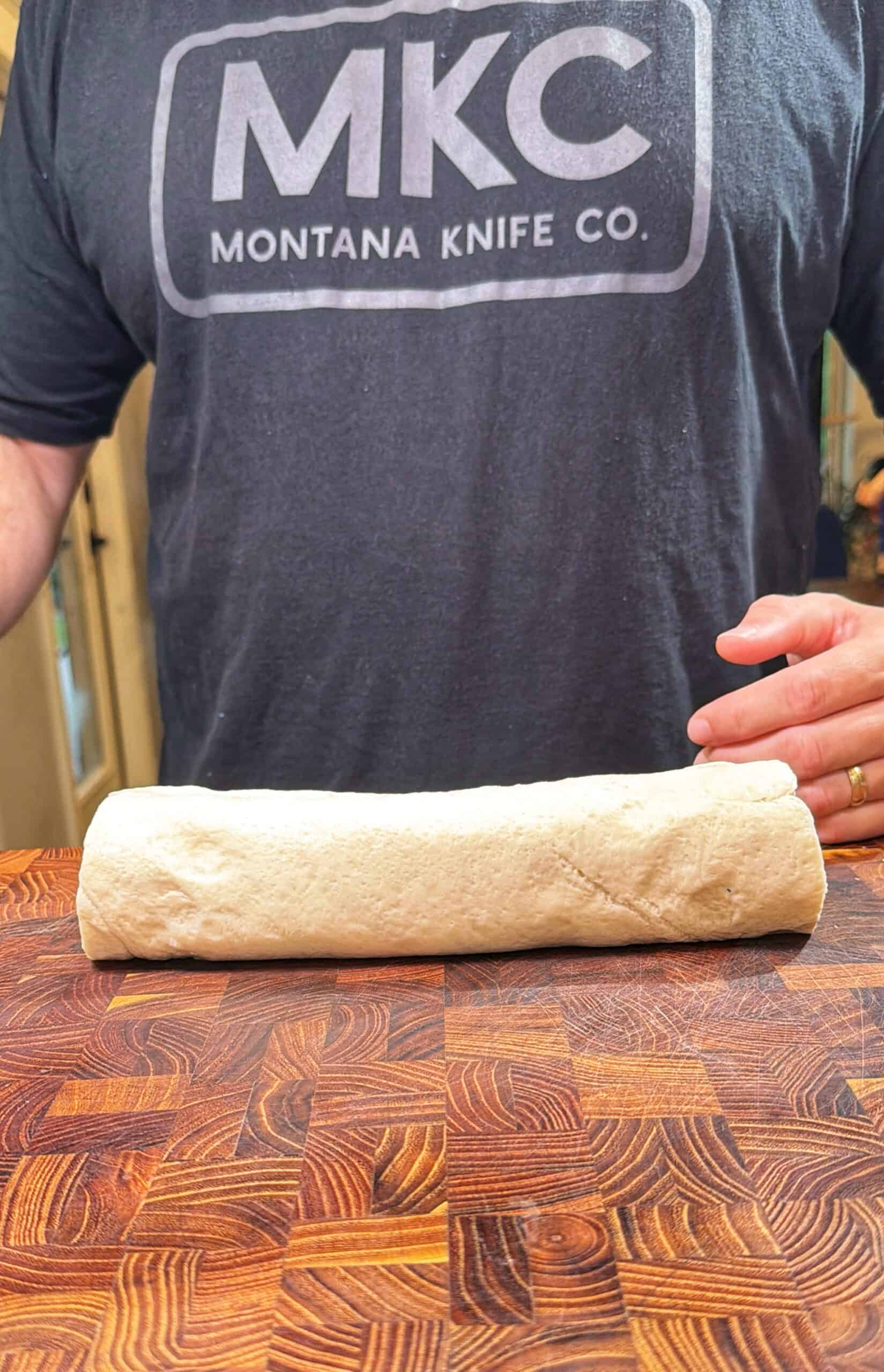 A person wearing a “Montana Knife Co.” t-shirt stands behind a rolled log of dough resting on a wooden cutting board.