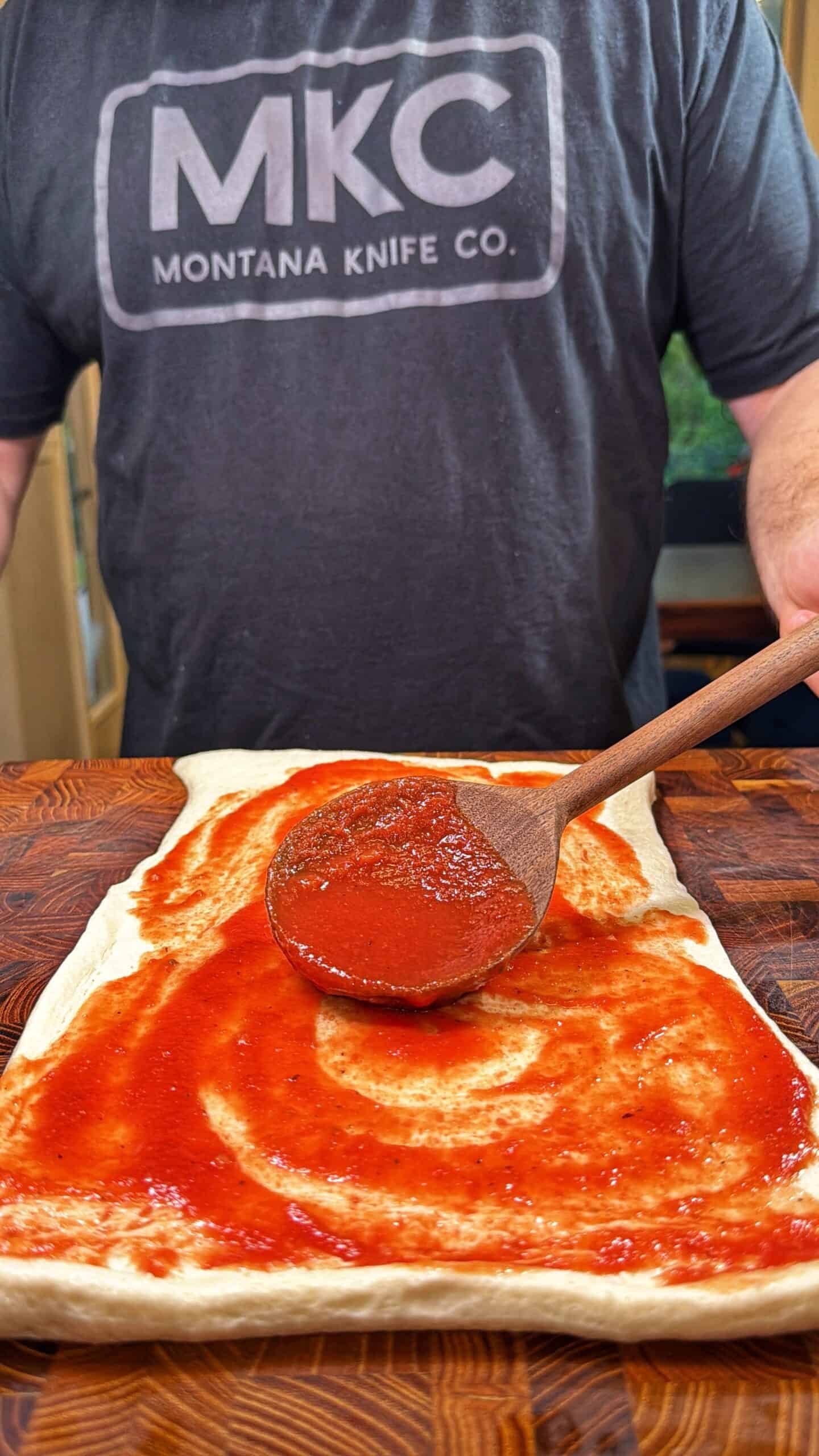 A person wearing an MKC Montana Knife Co. shirt spreads tomato sauce on pizza dough with a wooden spoon on a wooden surface.