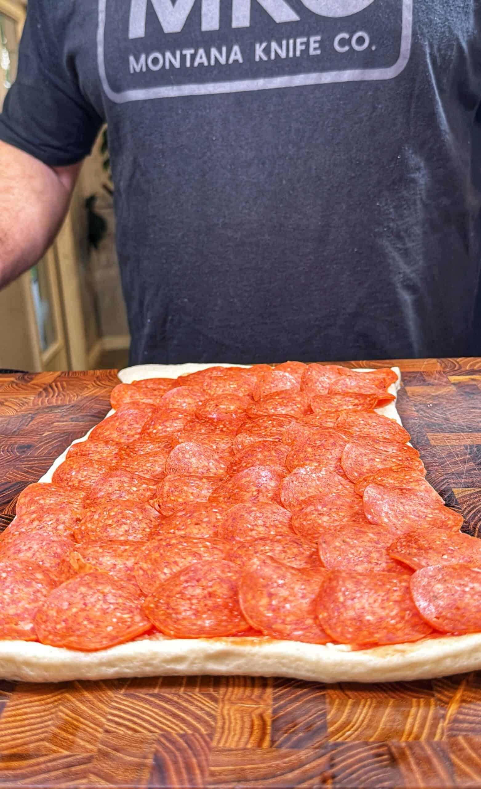 A person wearing a Montana Knife Co. shirt stands behind a rectangular sheet of dough covered with overlapping slices of pepperoni on a wooden surface.