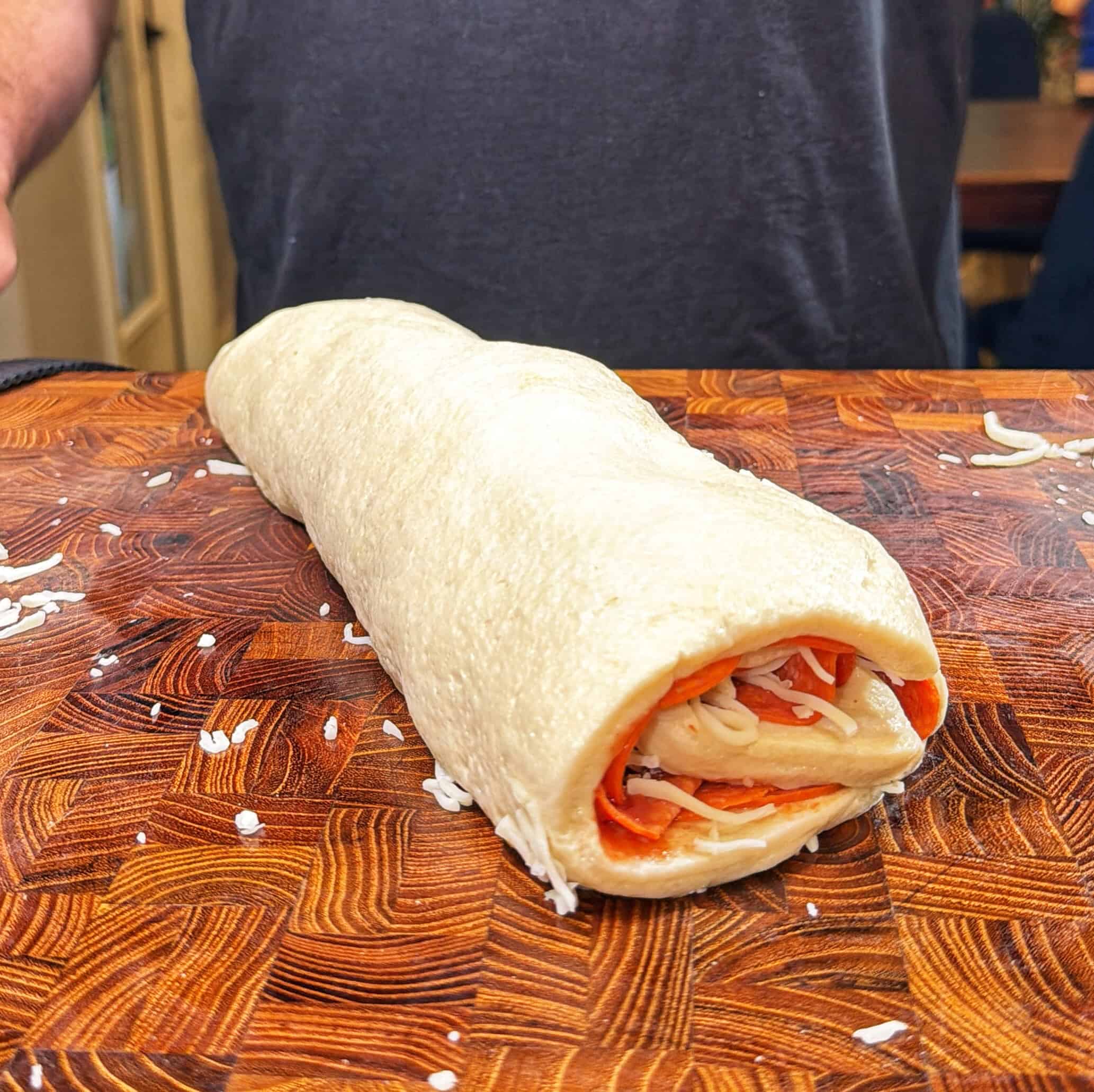 A rolled-up uncooked dough filled with cheese and pepperoni sits on a wooden cutting board, with a person in a dark shirt standing behind it.
