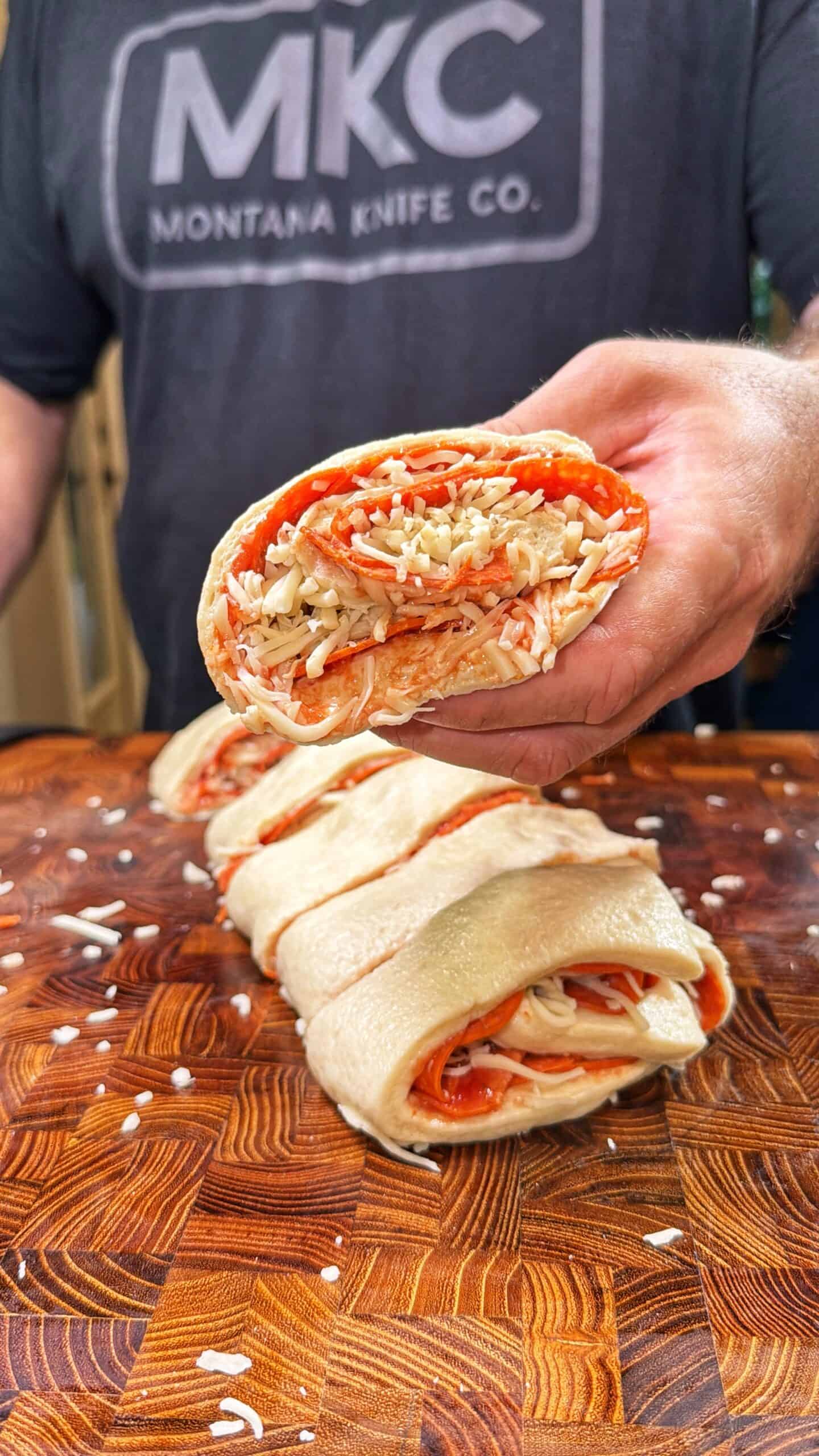 A person wearing a “Montana Knife Co.” shirt holds a sliced piece of a rolled dough stuffed with pepperoni and shredded cheese over a wooden cutting board with more slices.