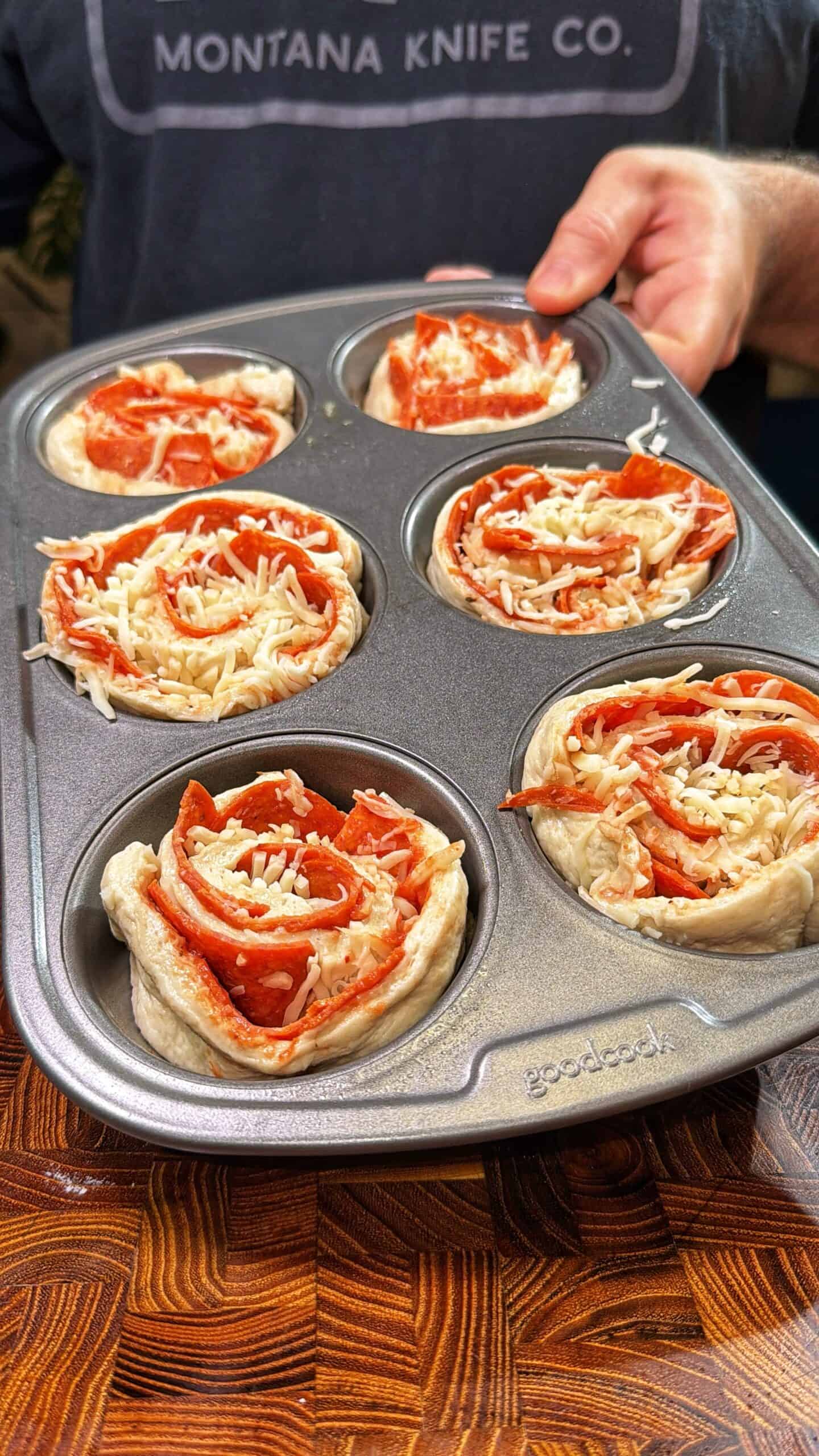 A person holds a muffin tin filled with unbaked pizza roll cups, each made of dough, pepperoni slices, and shredded cheese, ready to be baked. The person is wearing a Montana Knife Co. shirt.