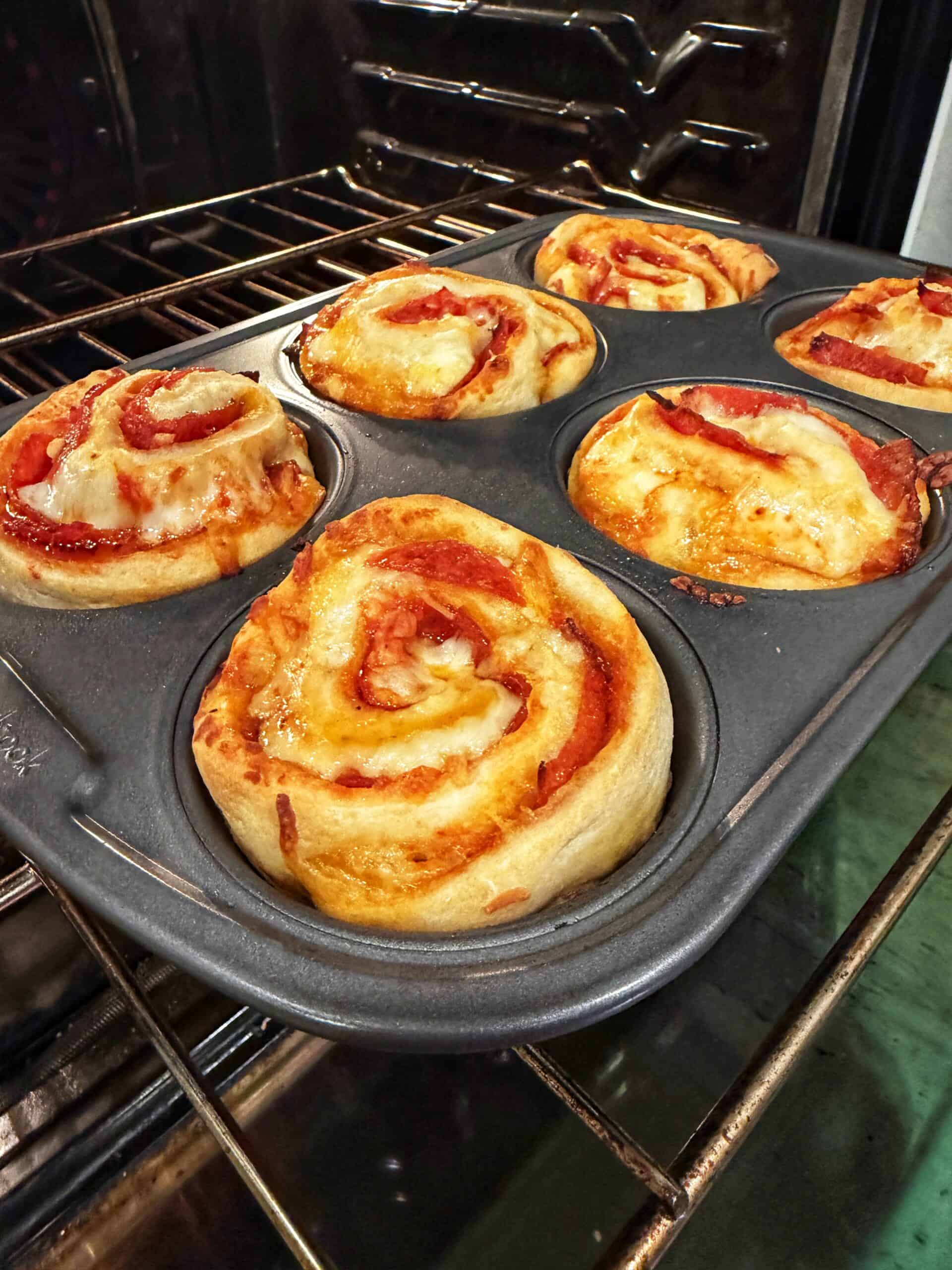 Six freshly baked pizza rolls in a muffin tin are resting on an oven rack. The rolls are golden brown with visible melted cheese and tomato sauce swirled in the dough.