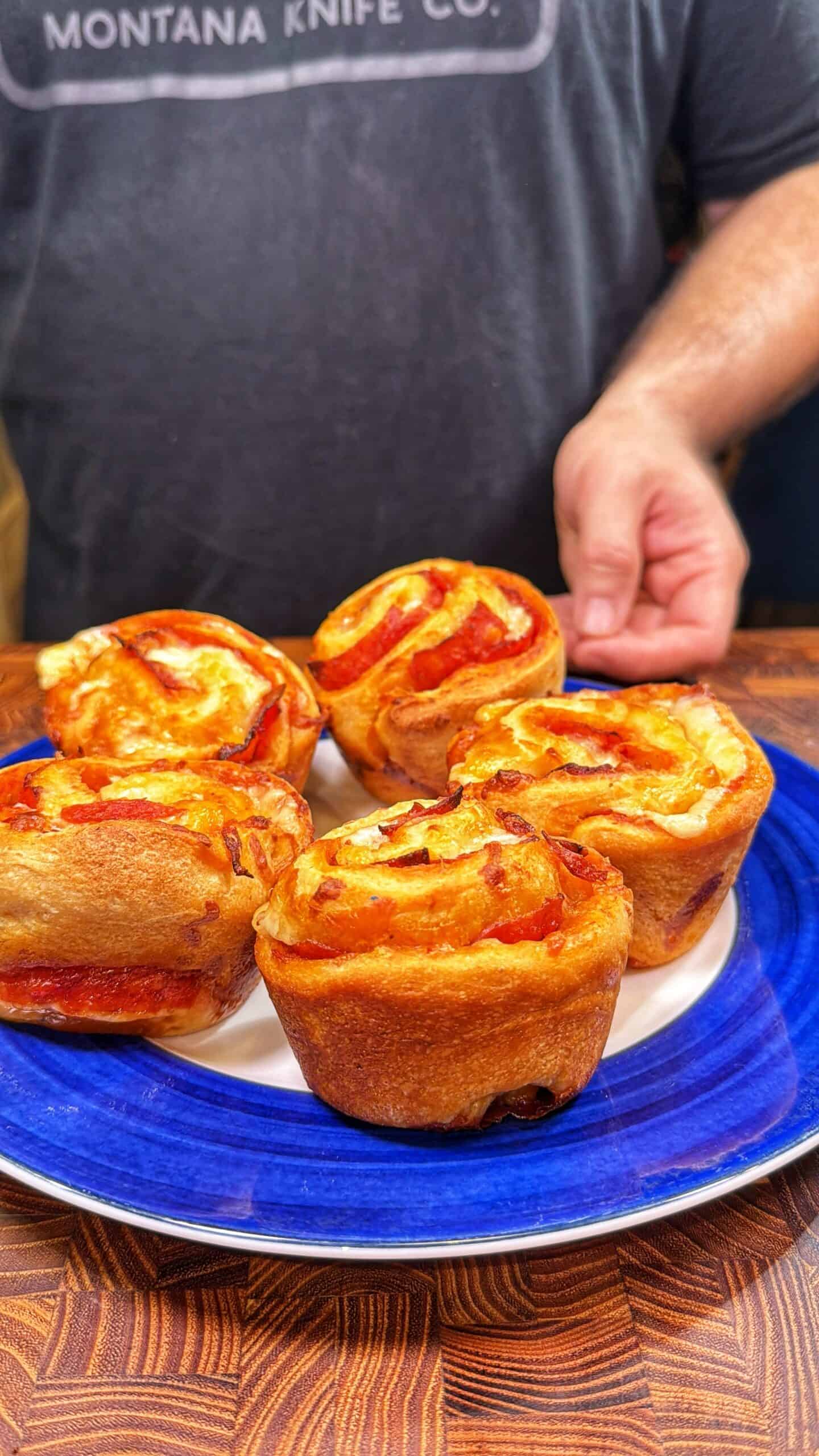 A person wearing a Montana Knife Co. shirt stands behind a plate with four golden, baked pizza muffins on a blue and white plate, reaching to pick one up.