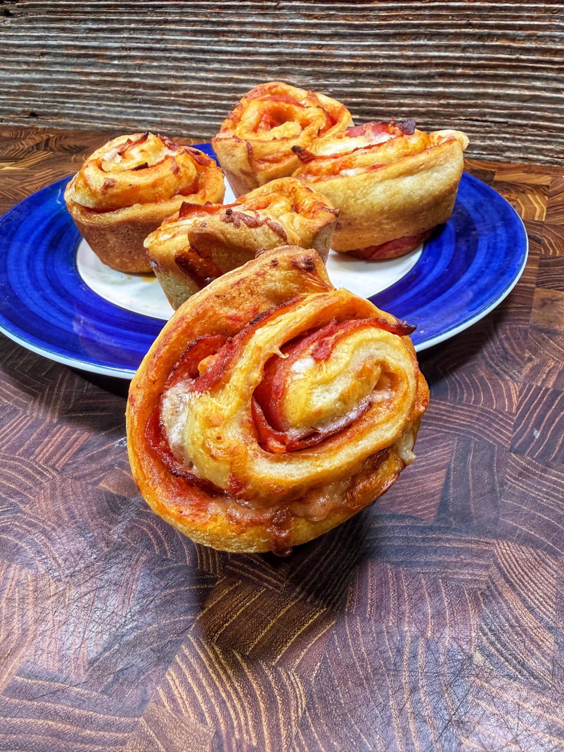 Four pizza rolls with visible layers of dough, cheese, and tomato sauce, are arranged on a blue plate, with one roll in the foreground on a wooden surface, and a rustic wood background behind them.