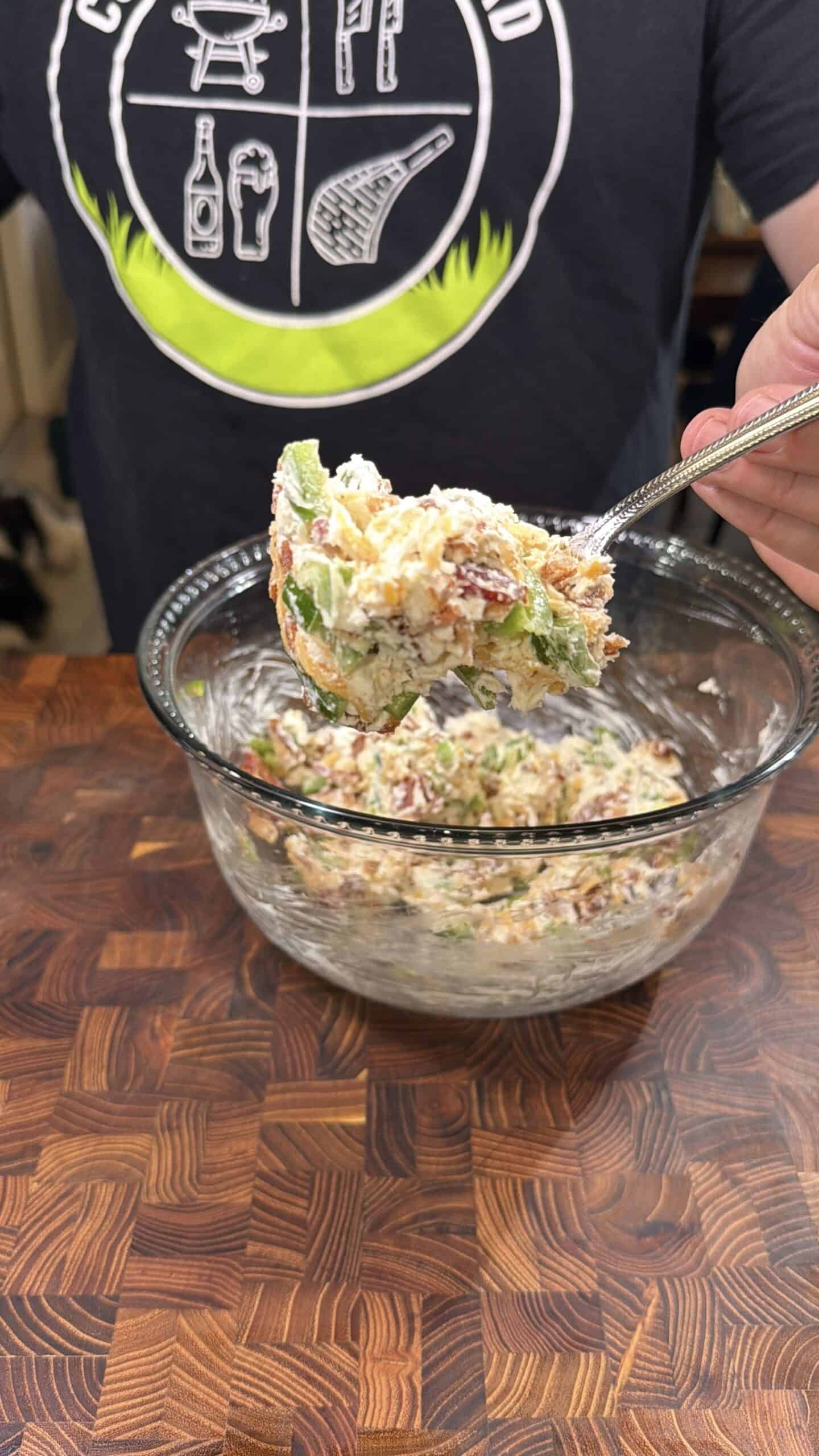A person holds a spoonful of a creamy salad mixture with vegetables and bacon over a glass bowl on a wooden countertop. The persons shirt shows grilling-themed graphics.