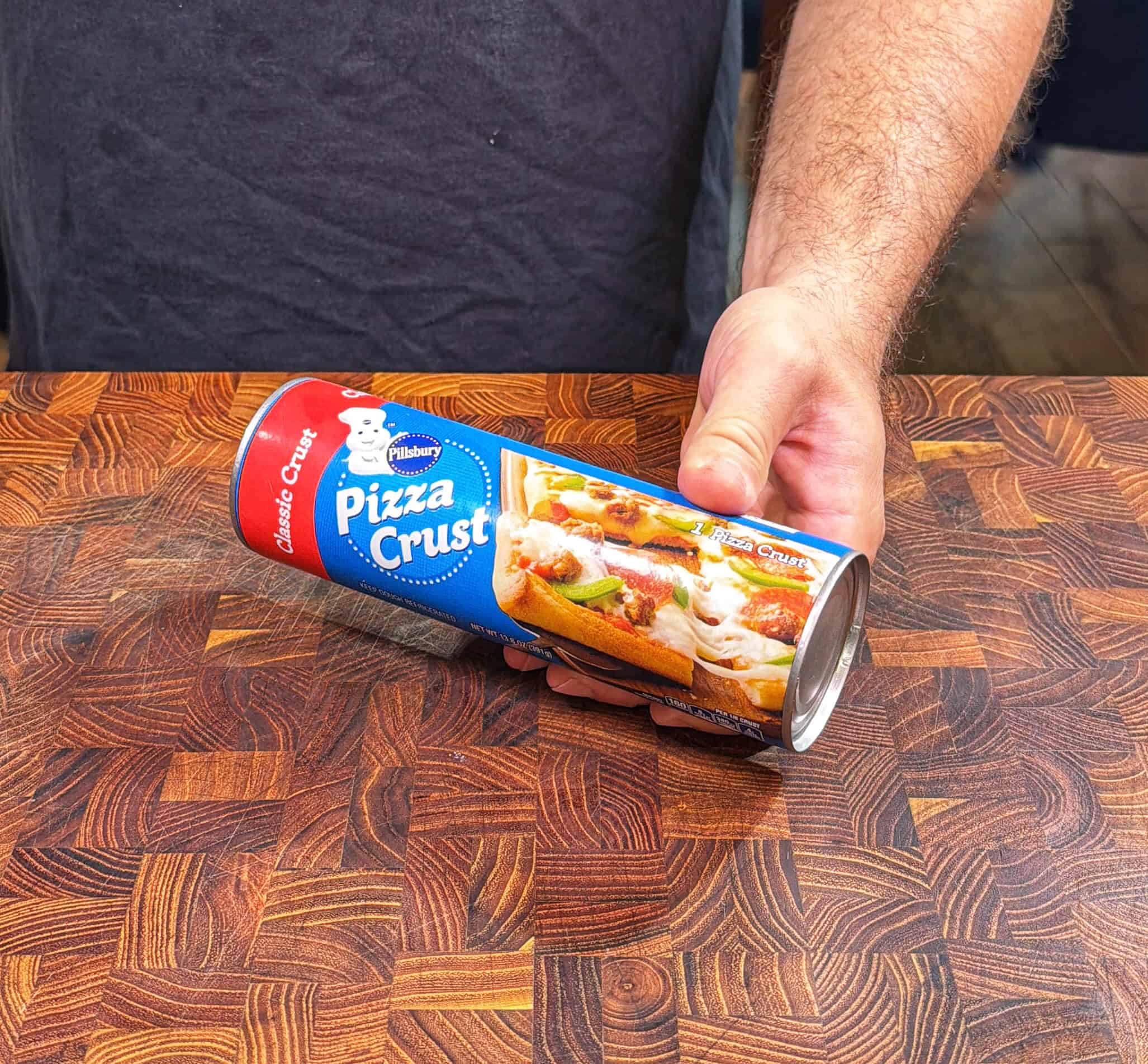 A person holds a can of Pillsbury Classic Crust Pizza Crust on a wooden countertop. The label shows an image of a baked pizza.