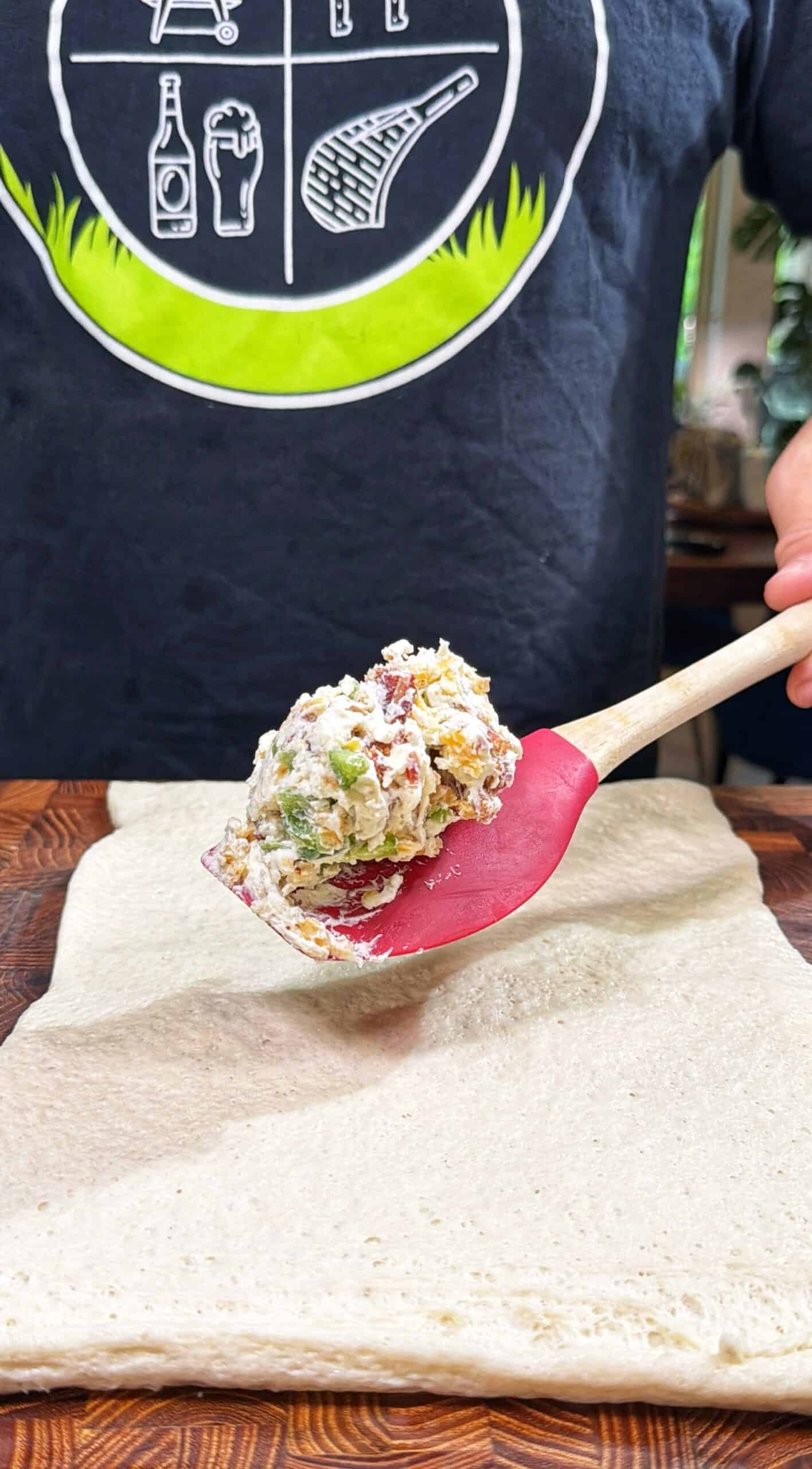 A person holds a red spatula with a scoop of creamy, colorful filling above a sheet of dough on a wooden surface. The person is wearing a dark shirt with a white and green graphic.