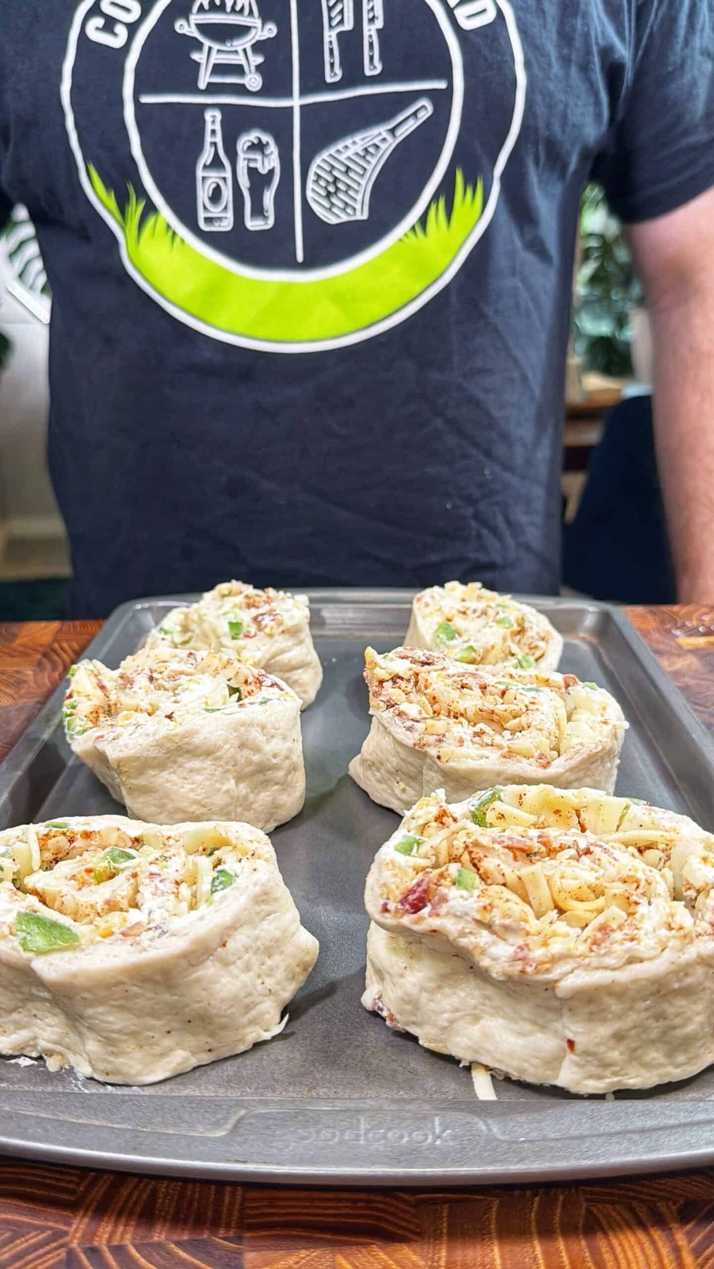 A person wearing a dark shirt with a food-themed graphic stands behind a baking tray filled with six uncooked savory pinwheel rolls on a wooden surface.
