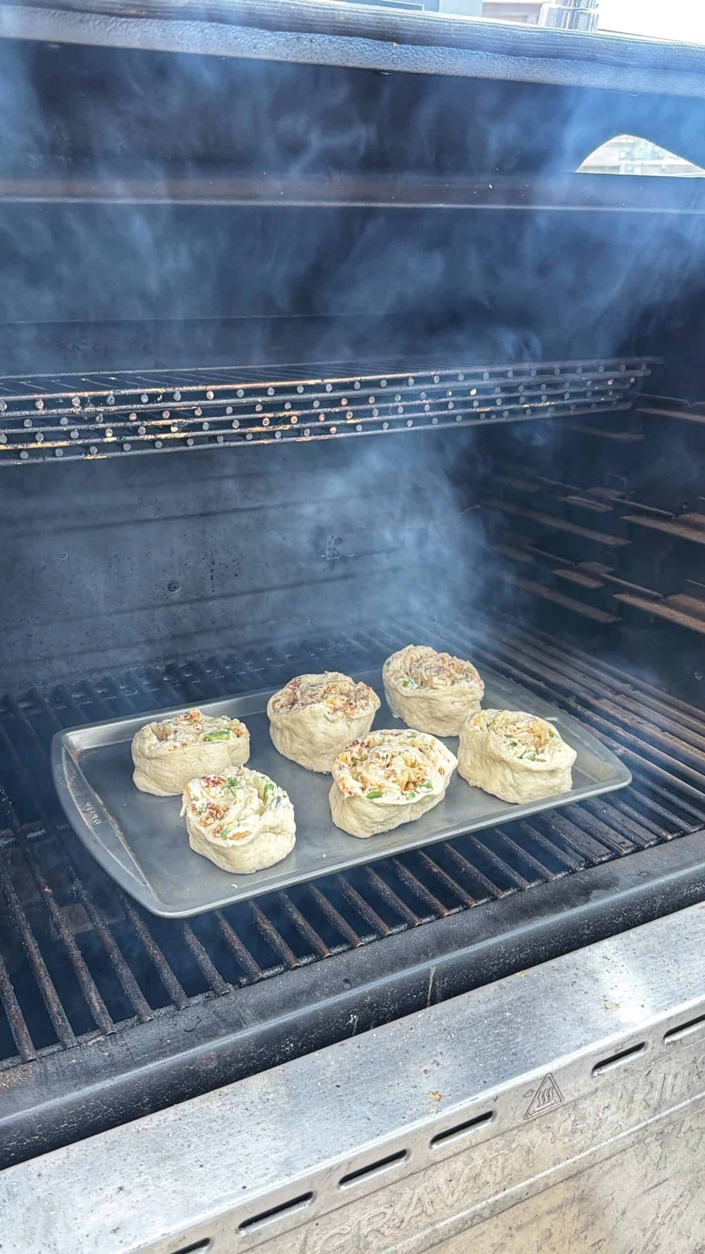 Six pieces of dough with toppings are baking on a tray inside a smoky outdoor grill, with the grill lid open and smoke rising around the food.