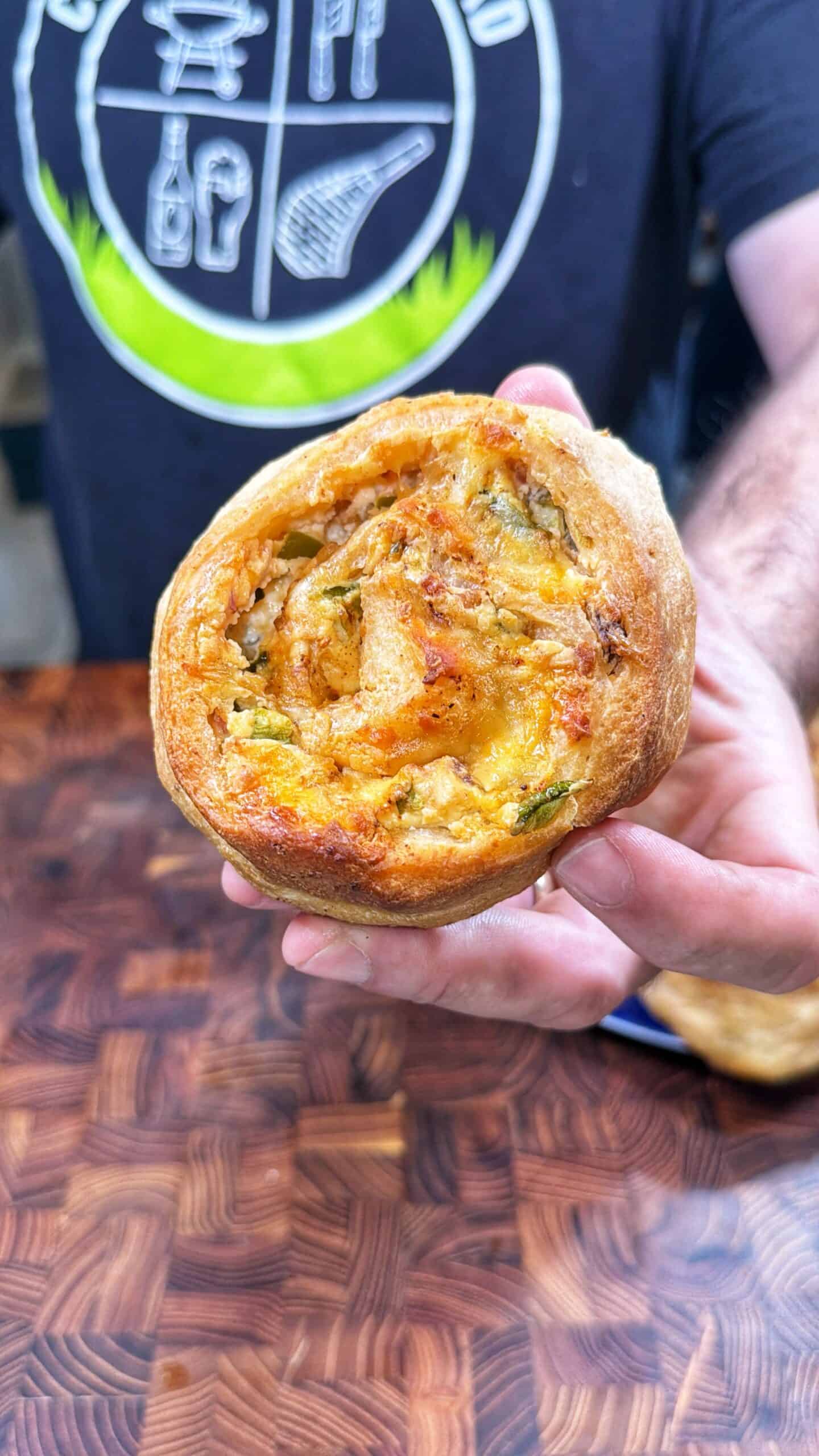 A person holds up a round, golden-brown savory pastry filled with melted cheese and vegetables. The person is wearing a black shirt with white graphics in the background.