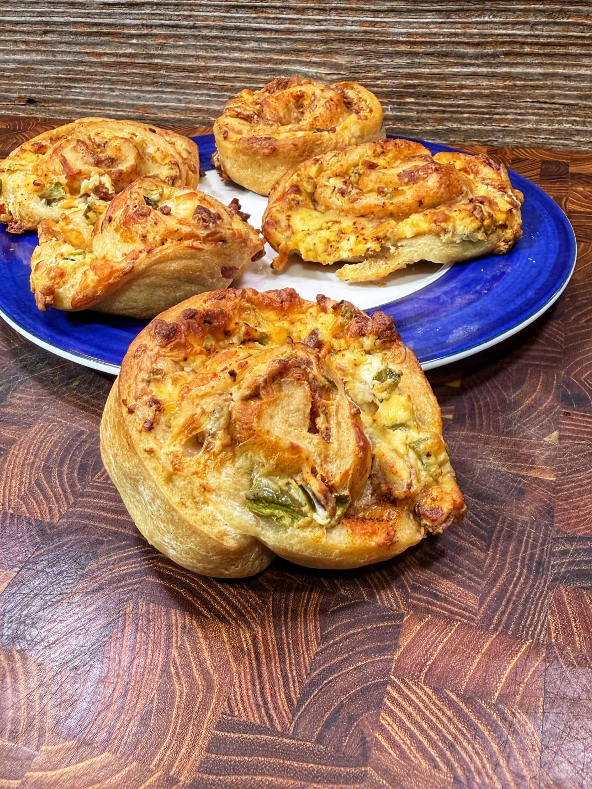 Four savory pinwheel pastries with cheese and vegetables are displayed on a blue plate and wooden surface, with one pastry placed in the foreground for emphasis.