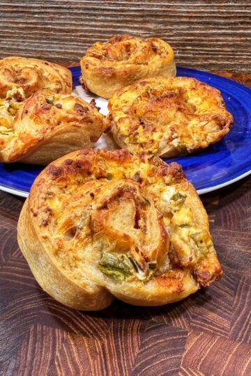 Four golden-brown, spiral-shaped savory rolls are displayed on a blue and white plate and a wooden surface. The rolls appear to contain cheese, vegetables, and possibly meat.