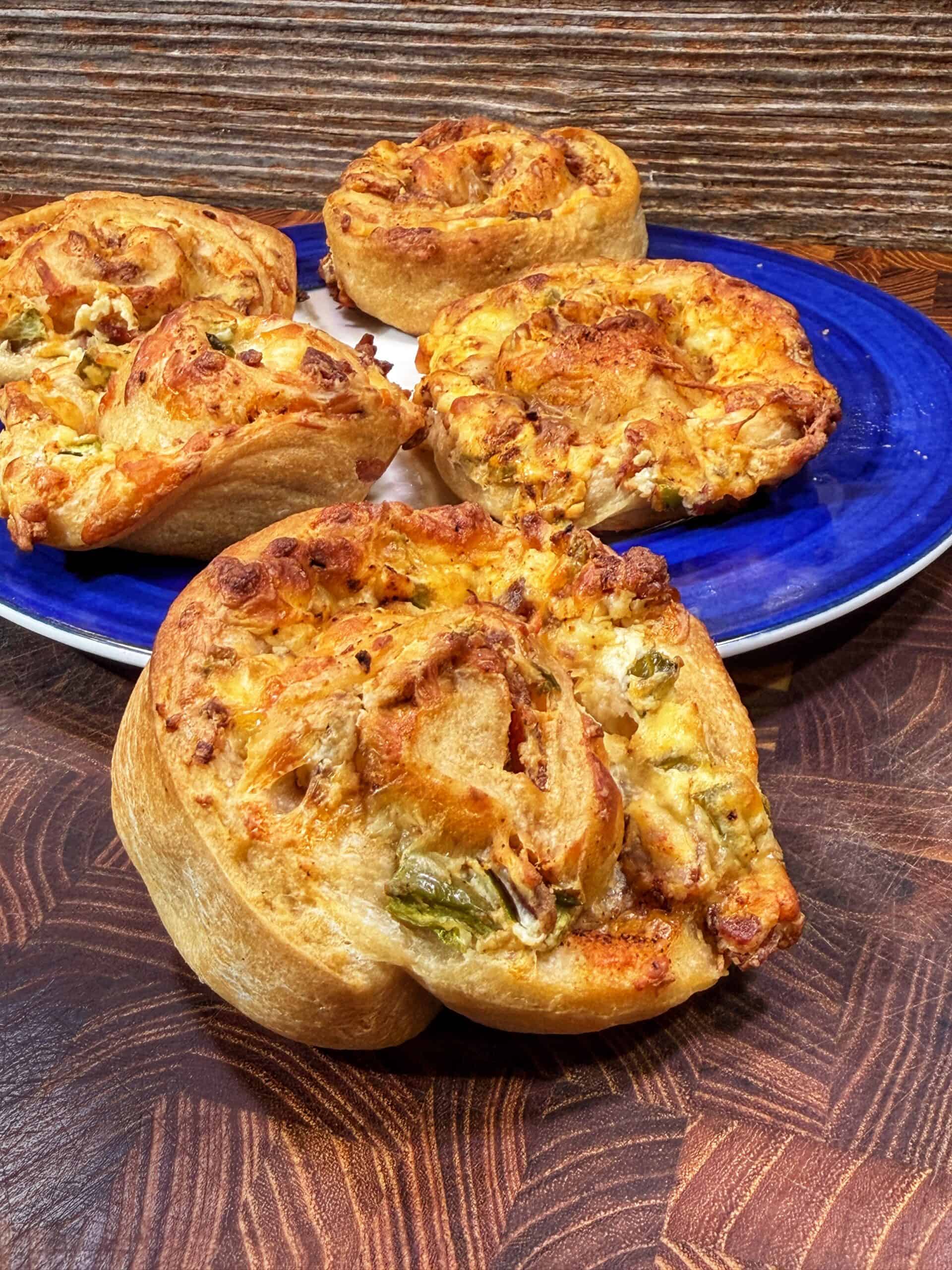 Four golden-brown, spiral-shaped savory rolls are displayed on a blue and white plate and a wooden surface. The rolls appear to contain cheese, vegetables, and possibly meat.