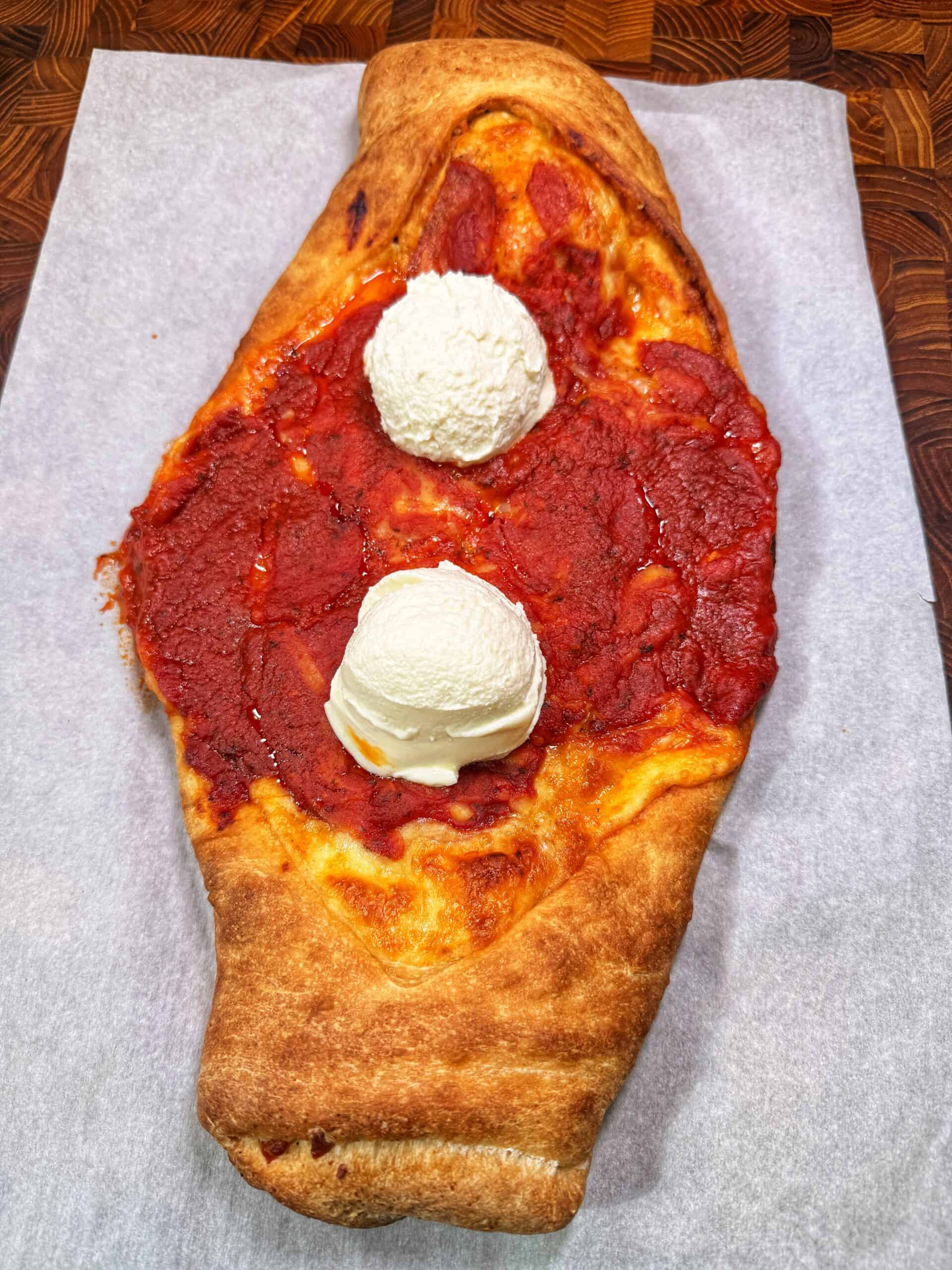 An oval-shaped pizza with a thick crust, topped with tomato sauce and two scoops of what appears to be vanilla ice cream, served on parchment paper on a wooden surface.