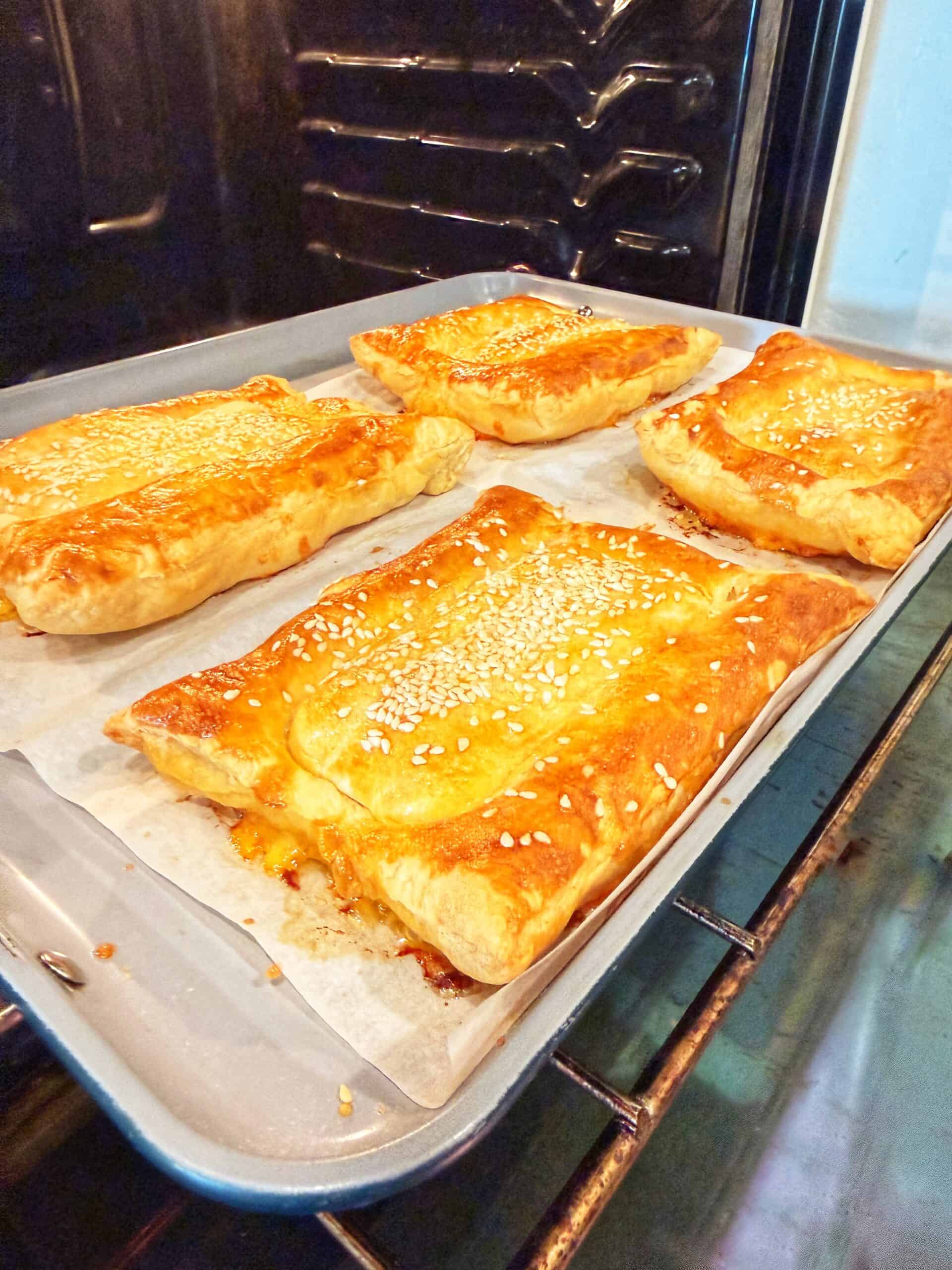 Four golden-brown puff pastry parcels topped with sesame seeds are baking on a parchment-lined tray inside an oven.