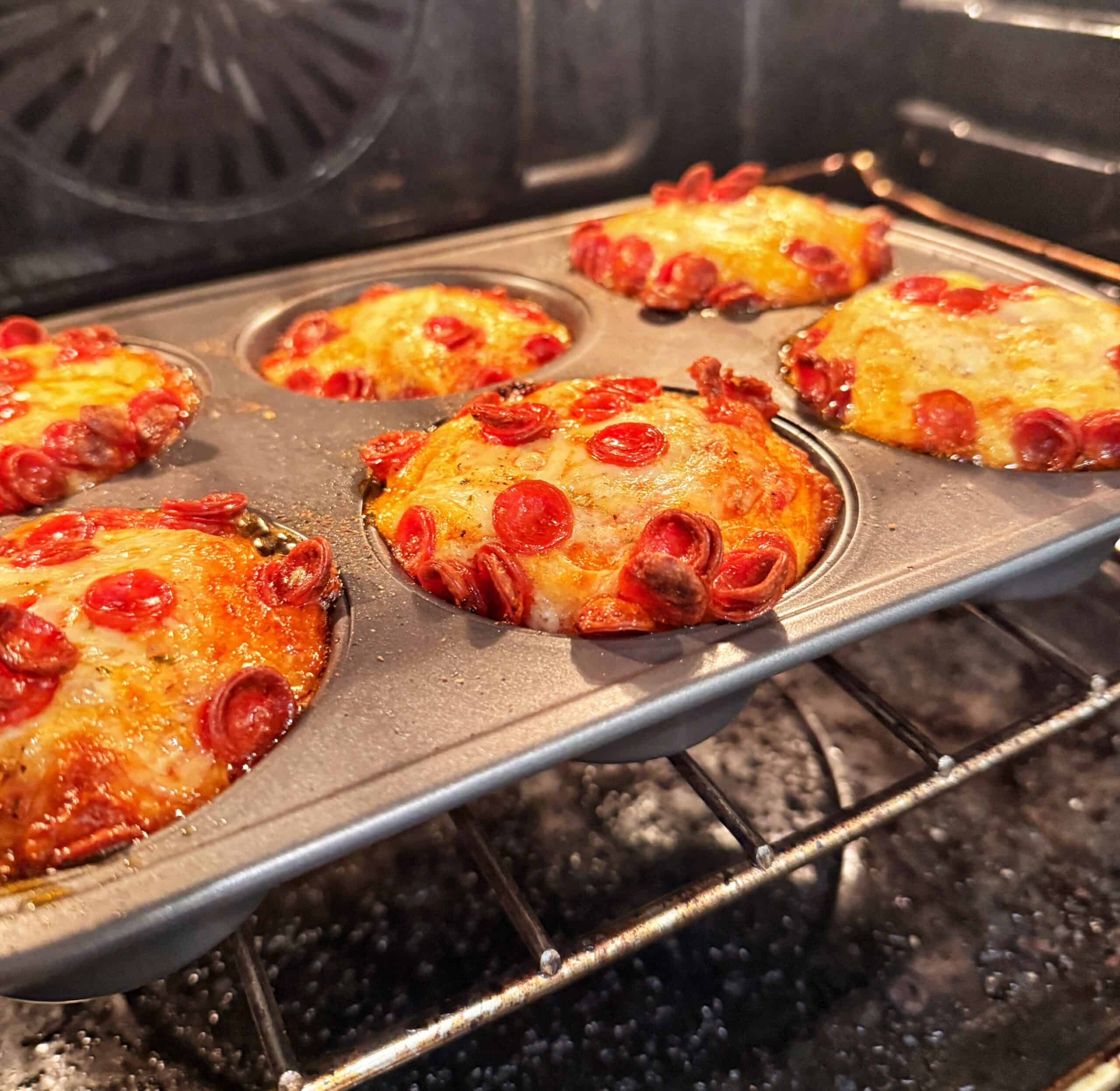 A muffin tin in an oven holds six baked mini pizzas topped with melted cheese and small pepperoni slices, with cheese bubbling and browning around the edges.
