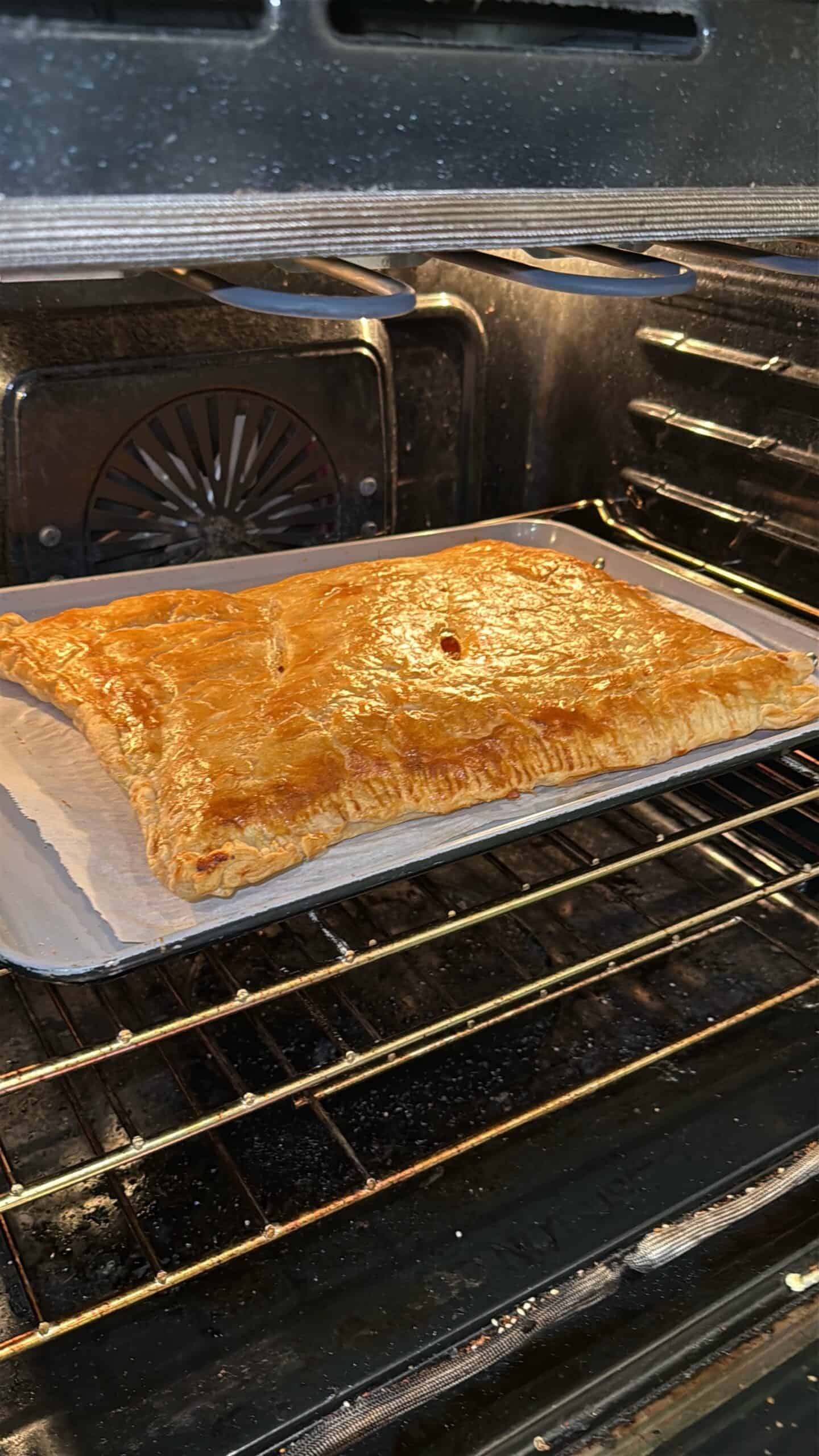 A large golden-brown pastry is baking on a parchment-lined tray inside an oven. The pastry appears flaky and puffed, with a shiny, crisp surface and a small vent hole in the center.
