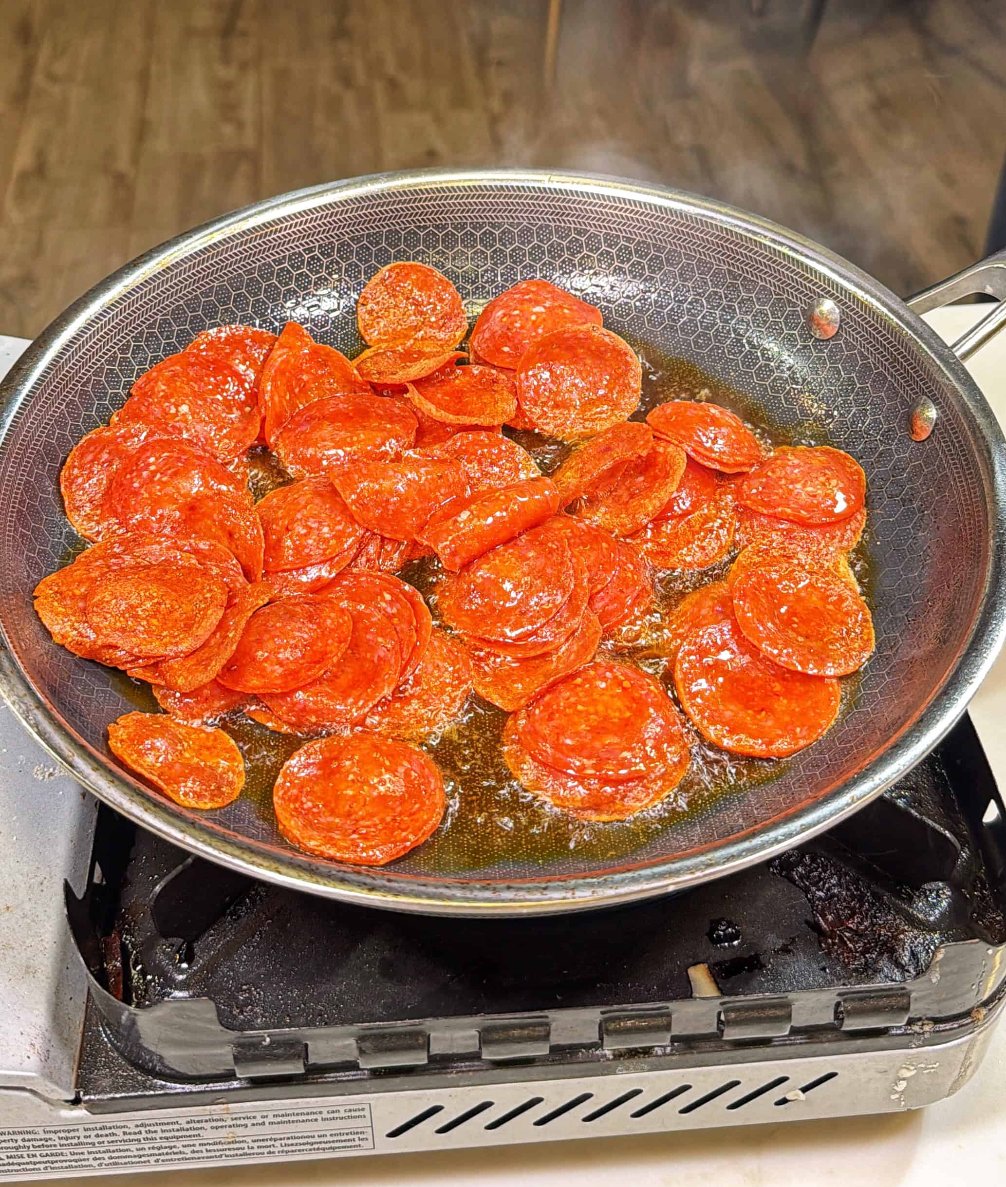 Sliced pepperoni is being cooked in oil in a frying pan on a portable stove. The pepperoni is sizzling and slightly curled at the edges. The stove sits on a countertop with a wood floor in the background.