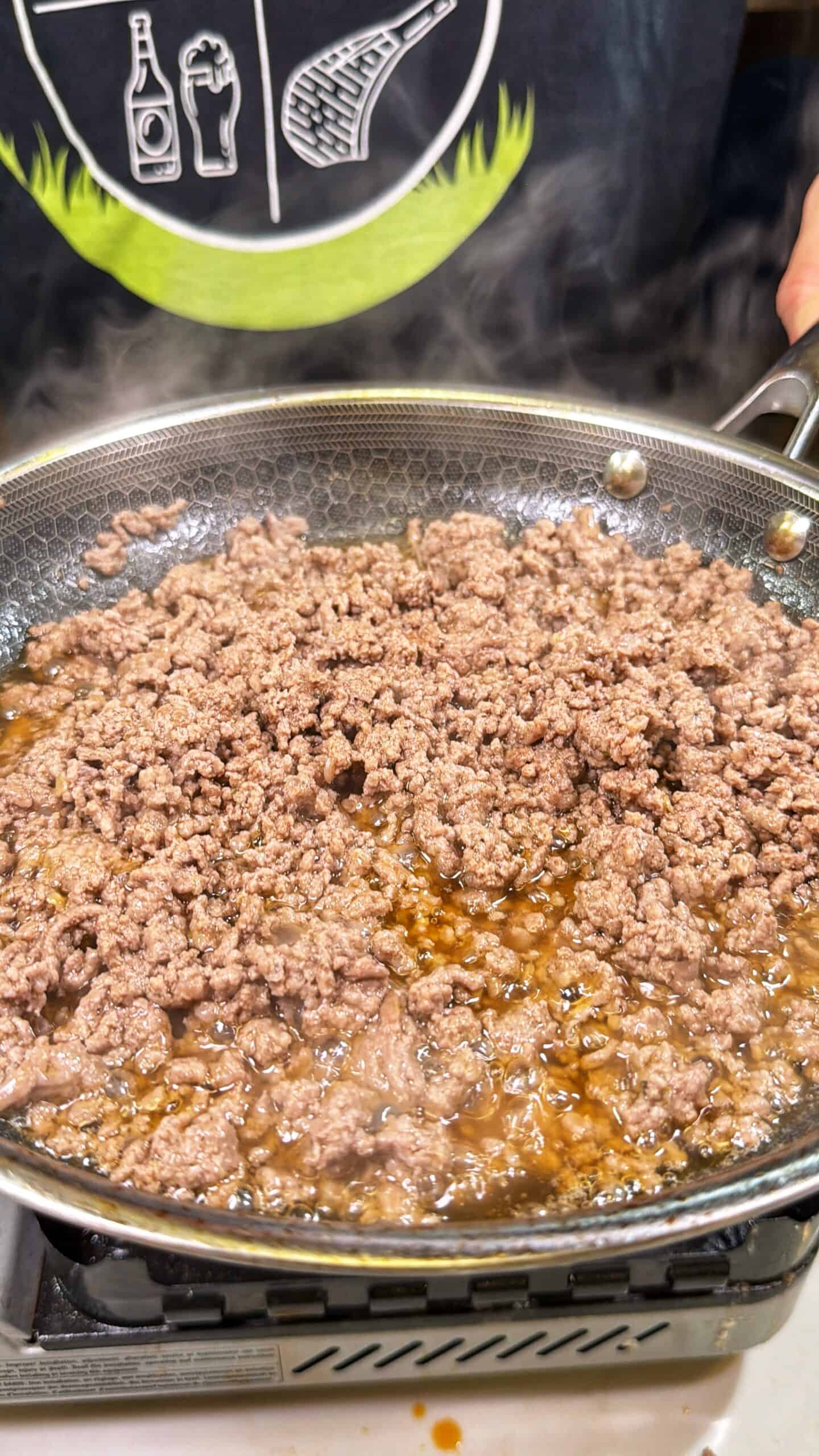 Ground beef is being browned in a frying pan on a stovetop. Steam rises from the meat as it cooks, and a person wearing a black shirt with food graphics is visible in the background.