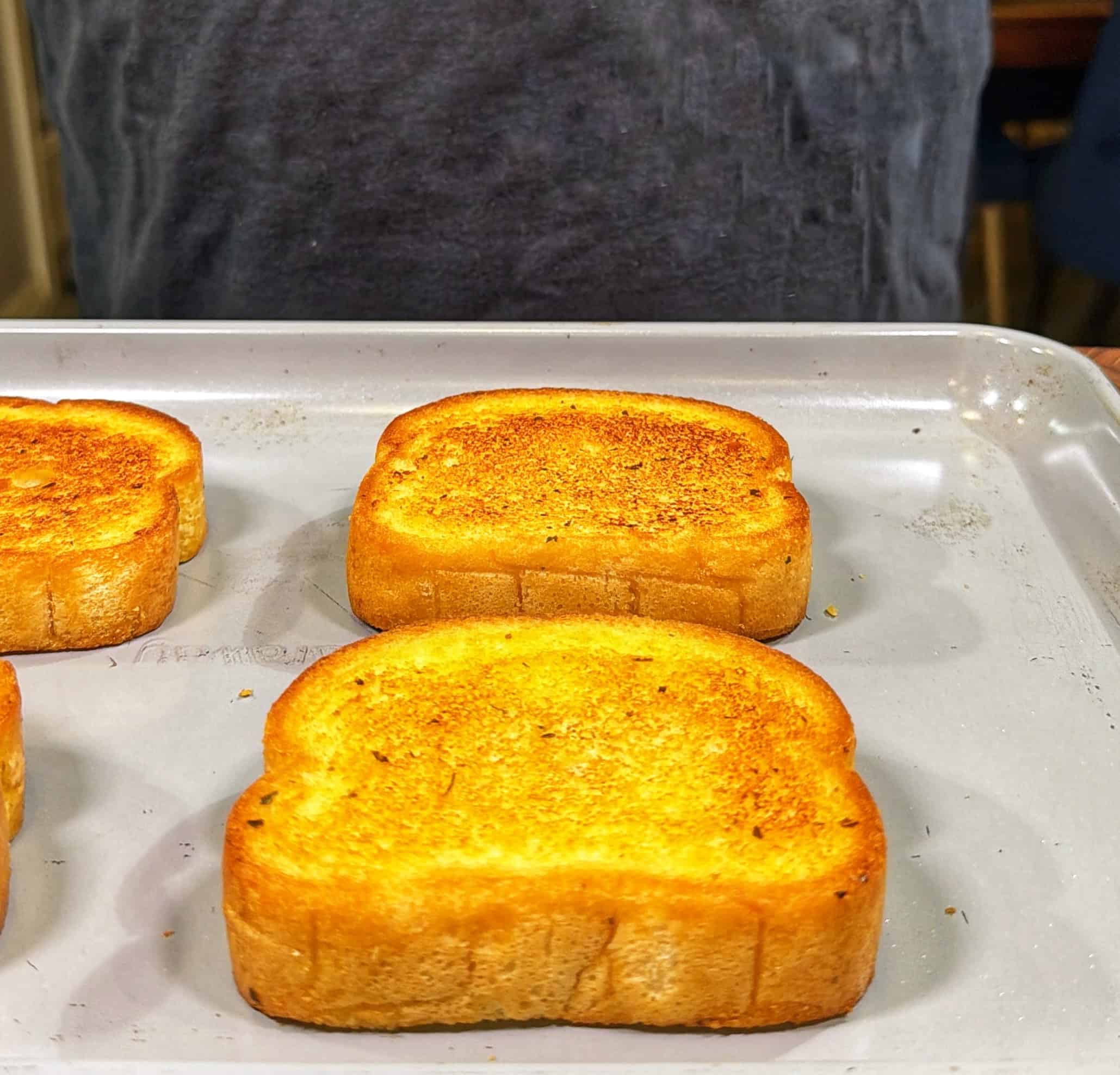 Three slices of golden-brown toasted bread sit on a baking sheet. A person in a gray shirt stands behind the tray, but only their torso is visible.