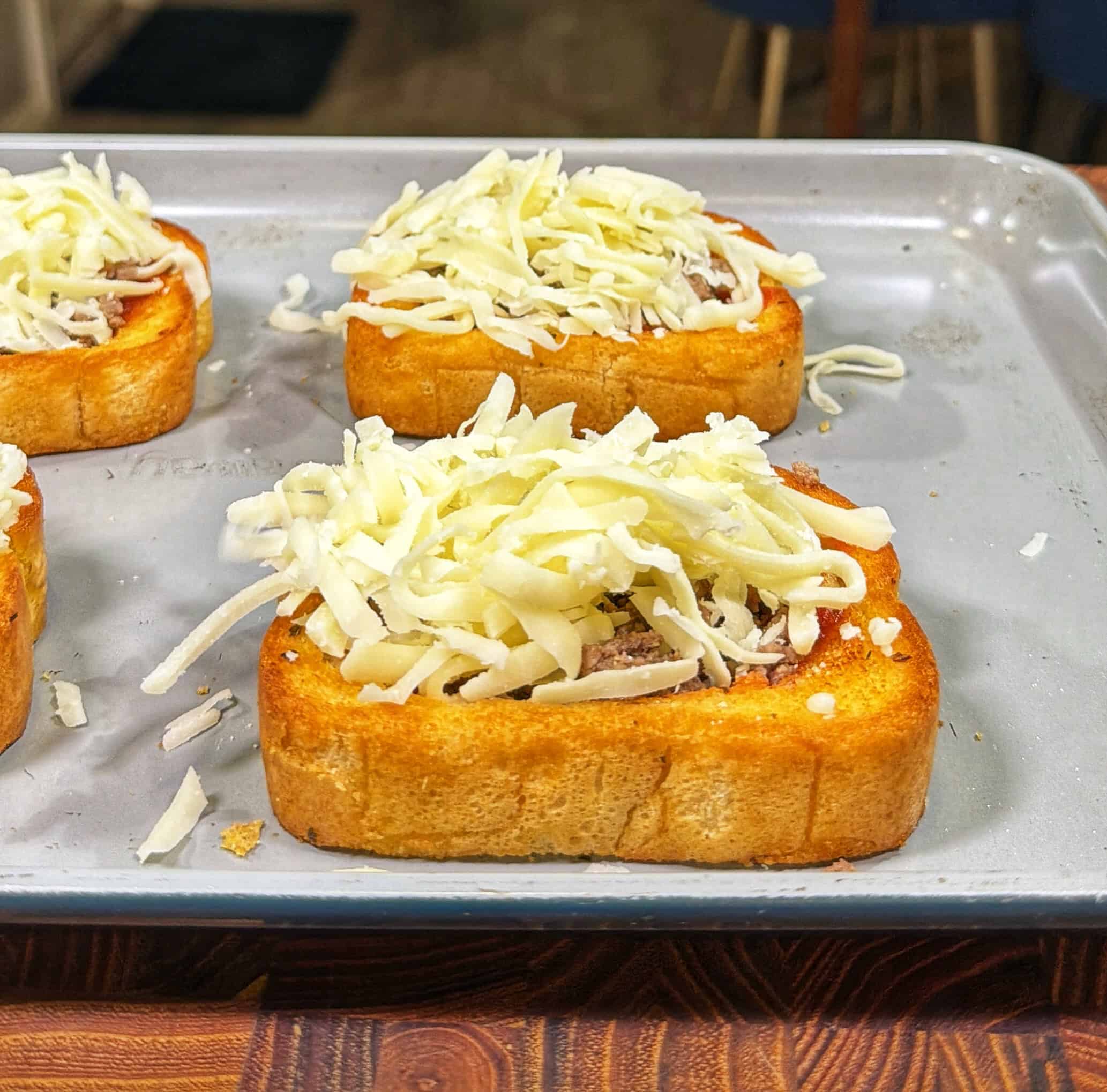 Slices of bread on a baking sheet topped with shredded cheese and other ingredients, prepared as mini pizzas before baking.