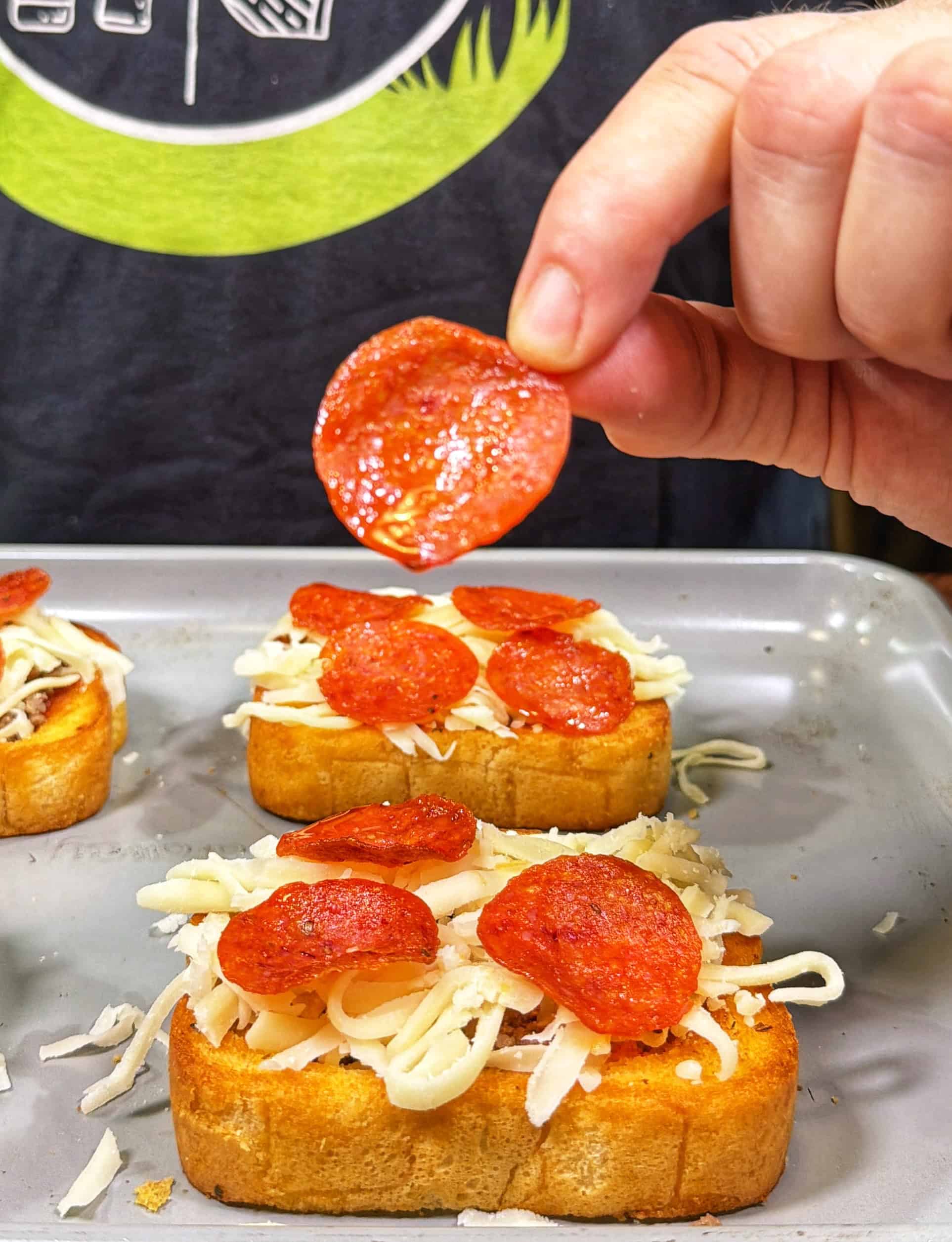 A hand places a slice of pepperoni onto a piece of garlic bread topped with shredded cheese and other pepperoni slices, preparing mini pizzas on a baking sheet.