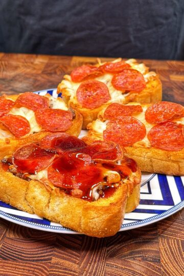 Three slices of thick bread topped with melted cheese and pepperoni are arranged on a decorative plate, with one slice featuring a glossy, reddish sauce. The plate sits on a wooden surface.