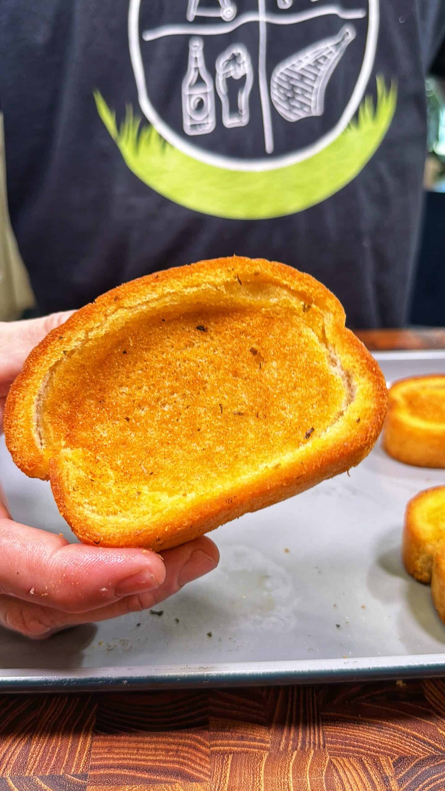 A hand holds a slice of golden-brown toasted bread over a baking tray, with more slices in the background. The person is wearing a dark shirt with white food-themed graphics.