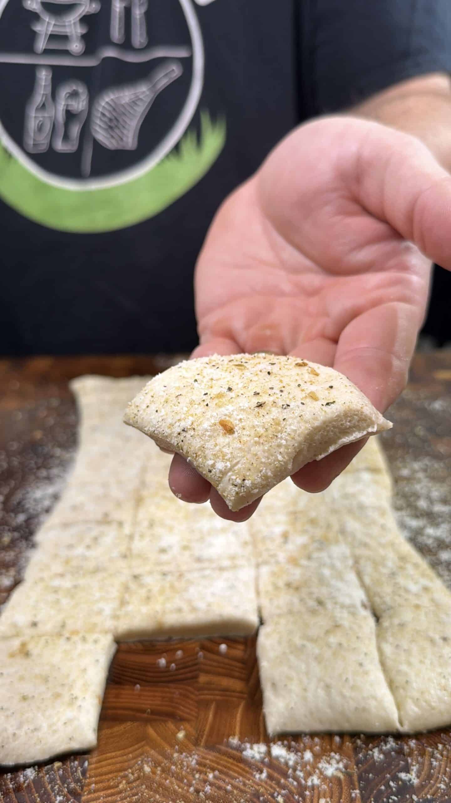 A hand holds a square piece of seasoned, uncooked dough above a wooden surface, with more dough pieces and flour in the background. The person is wearing a dark shirt with graphic illustrations.