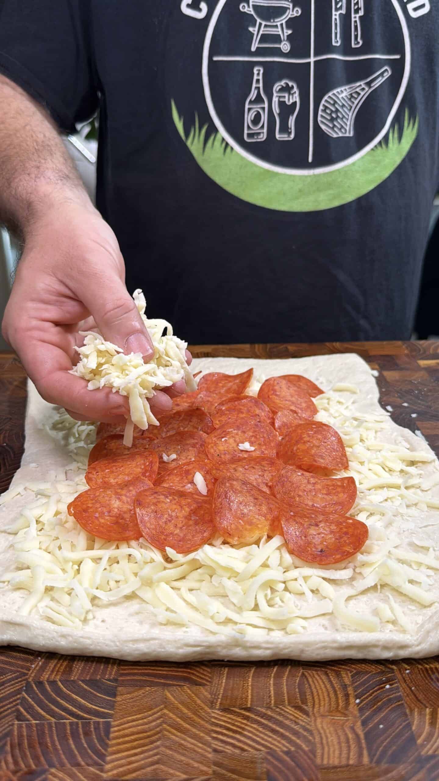 A person spreads shredded cheese over a layer of pepperoni slices on rectangular pizza dough, preparing a pizza on a wooden surface. The person is wearing a black shirt with white graphics.