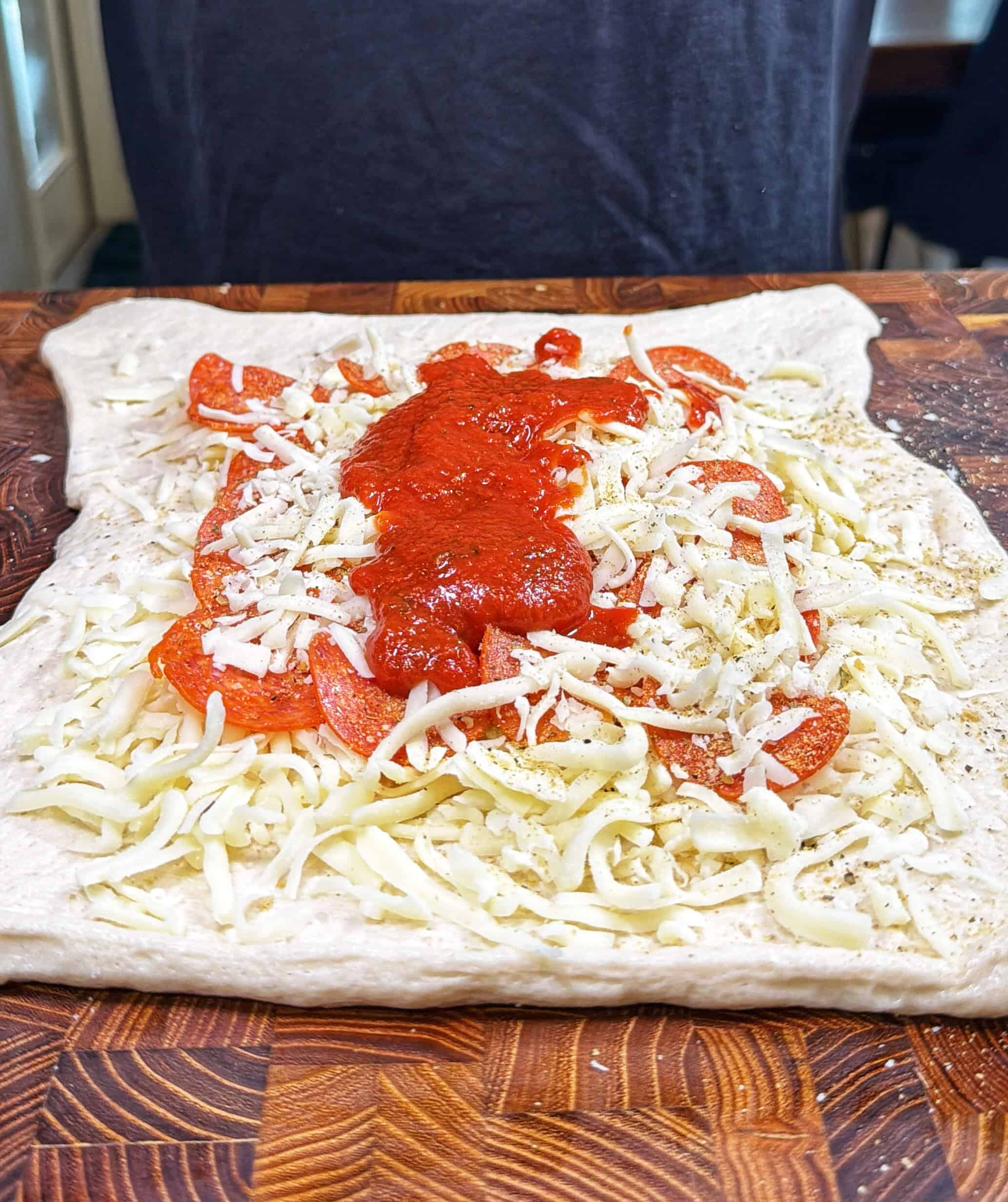 A rectangular piece of pizza dough topped with shredded cheese, pepperoni slices, Italian seasoning, and a layer of tomato sauce sits on a wooden cutting board. A person stands in the background.
