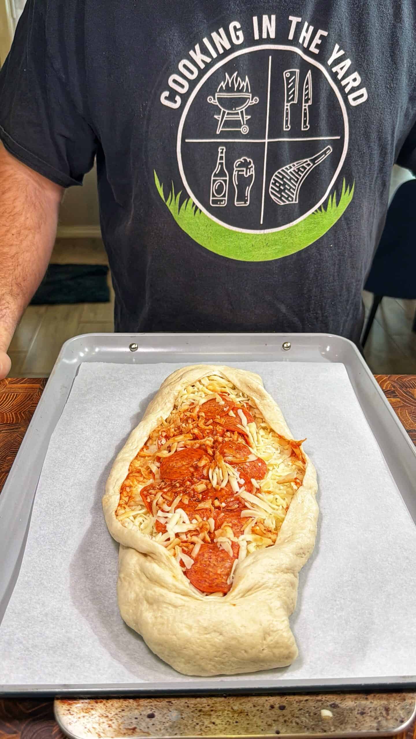 A person wearing a Cooking in the Yard shirt stands behind an unbaked, boat-shaped pepperoni pizza on a baking tray, ready to be put in the oven.