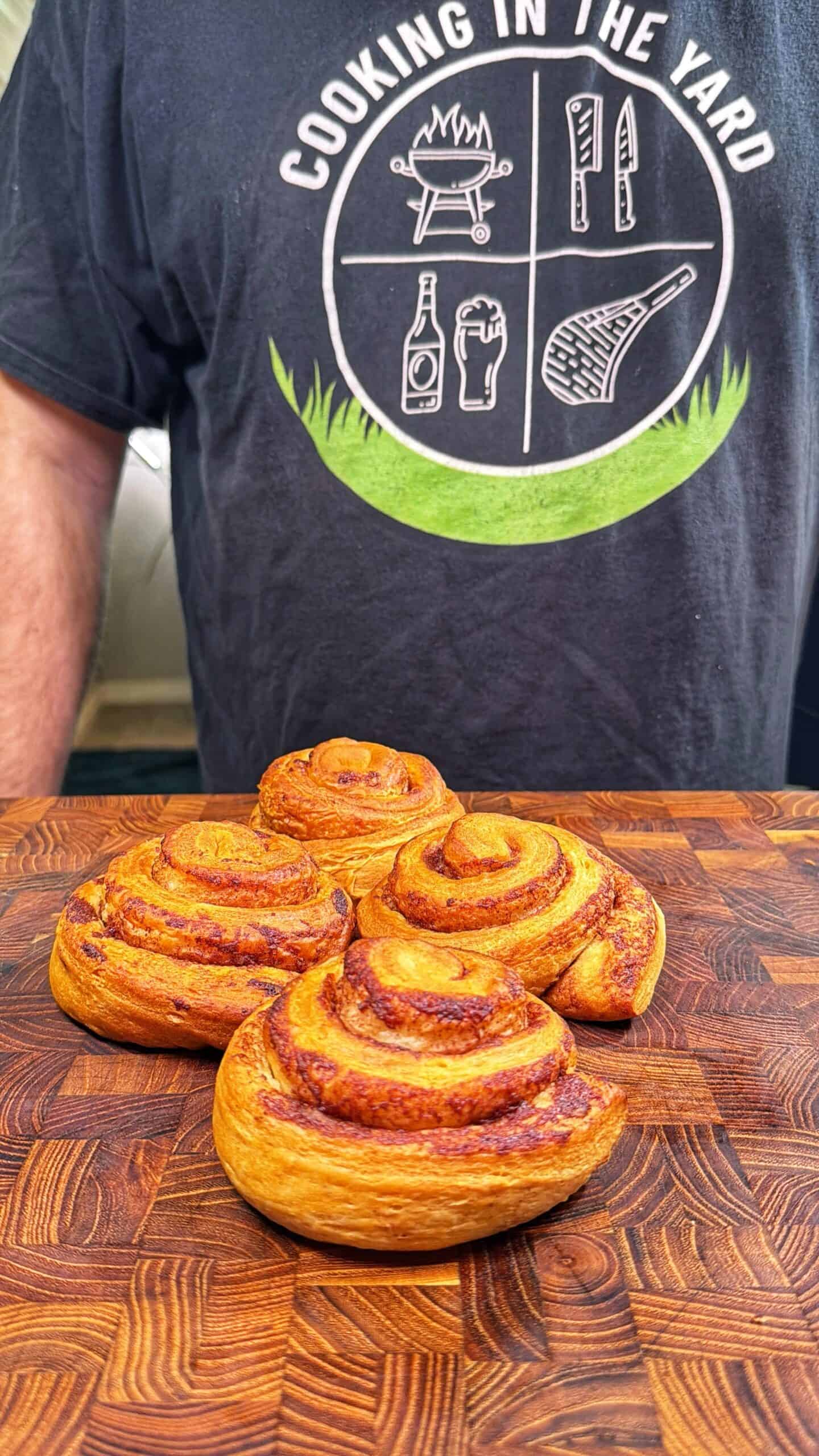 Four cinnamon rolls sit on a wooden cutting board. In the background, a person wears a black shirt that says COOKING IN THE YARD with grill and food icons printed on it.