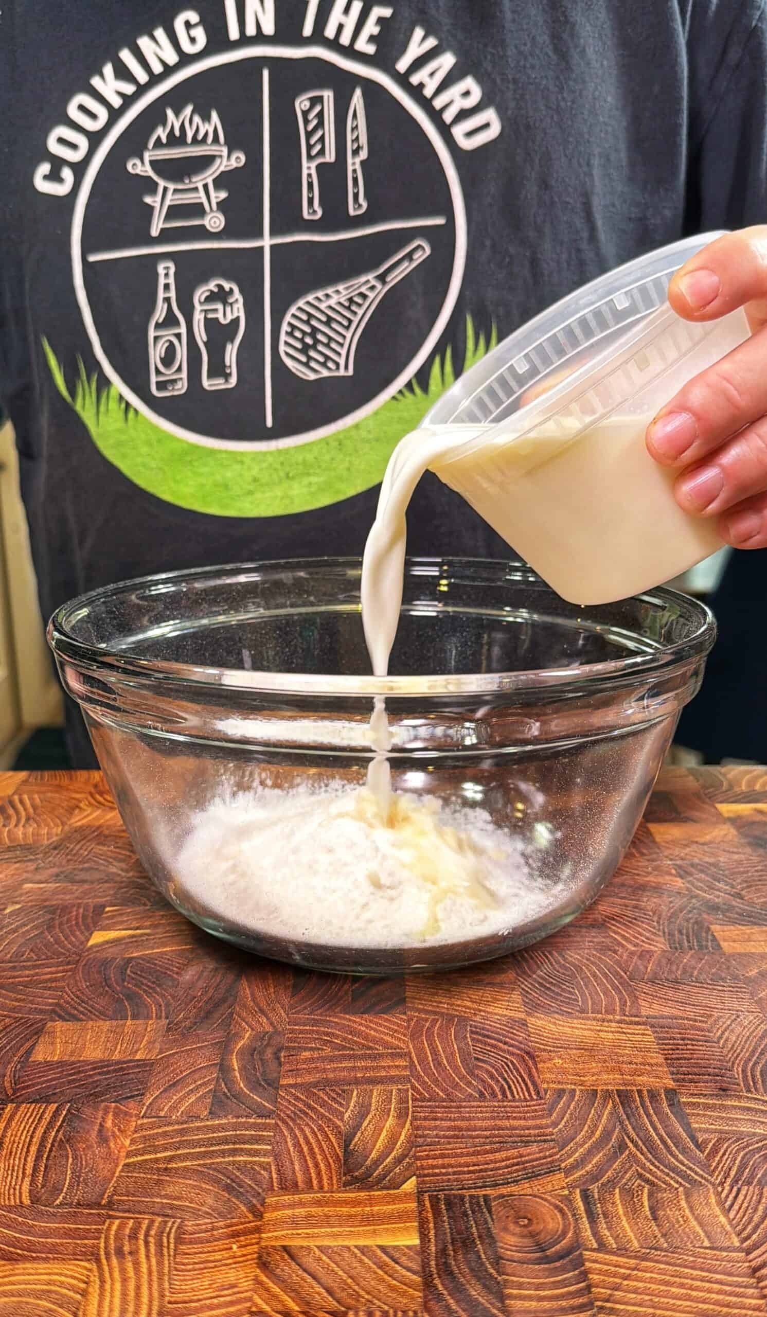 A person pours milk from a plastic container into a glass bowl of flour on a wooden countertop. The person is wearing a gray shirt with cooking-themed graphics and the words COOKING IN THE YARD.