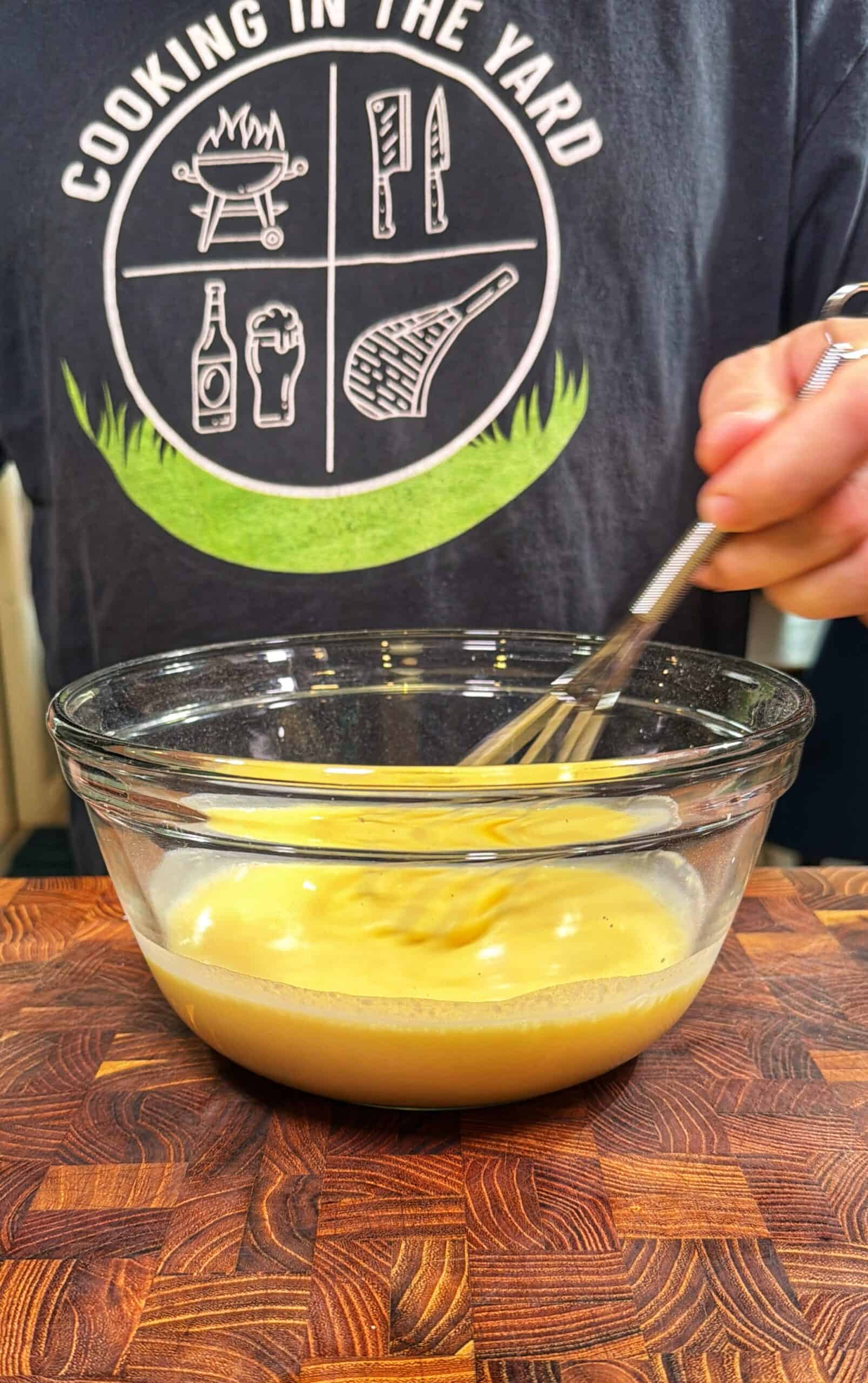 A person wearing a “Cooking in the Yard” shirt whisks a yellow mixture in a glass bowl on a wooden surface.