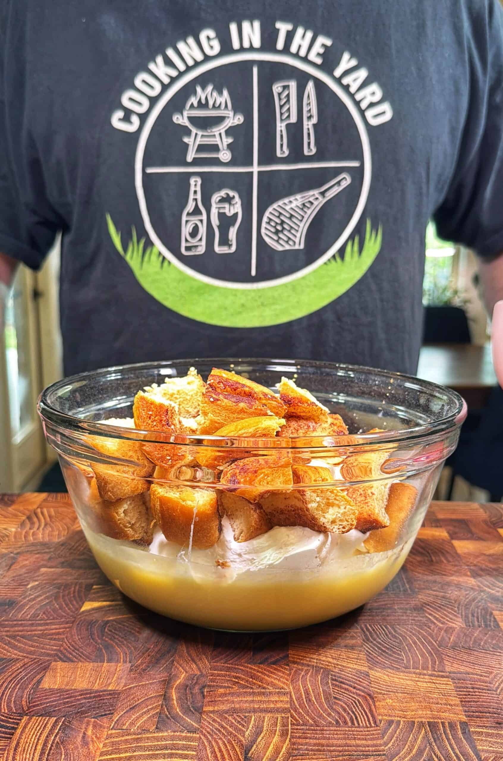 A glass bowl filled with torn pieces of bread soaking in liquid sits on a wooden surface. Behind it, a person wearing a Cooking in the Yard t-shirt is visible from the shoulders down.