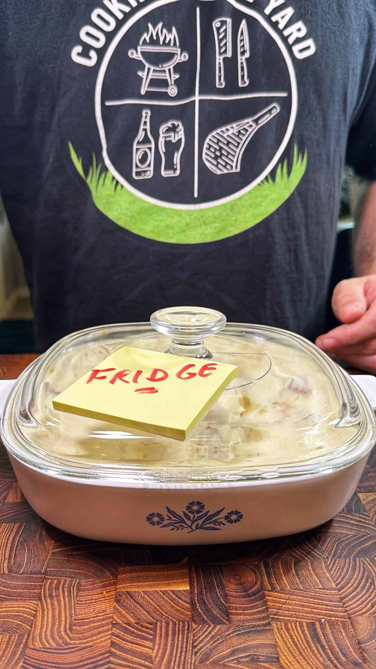 A glass casserole dish with a lid and a yellow sticky note labeled FRIDGE sits on a wooden surface. Behind it, a person wearing a Cooking Is My Card t-shirt is visible from the chest down.