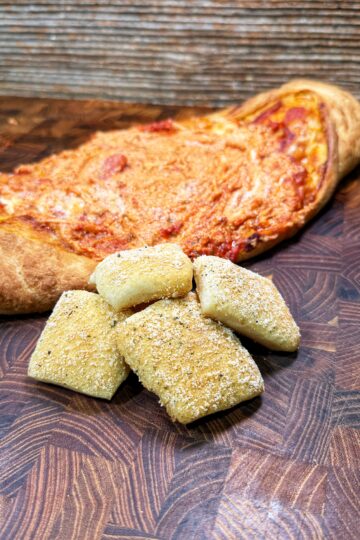A golden brown calzone sits on a wooden cutting board, with several rectangular pieces of bread dusted with seasoning placed in front of it.