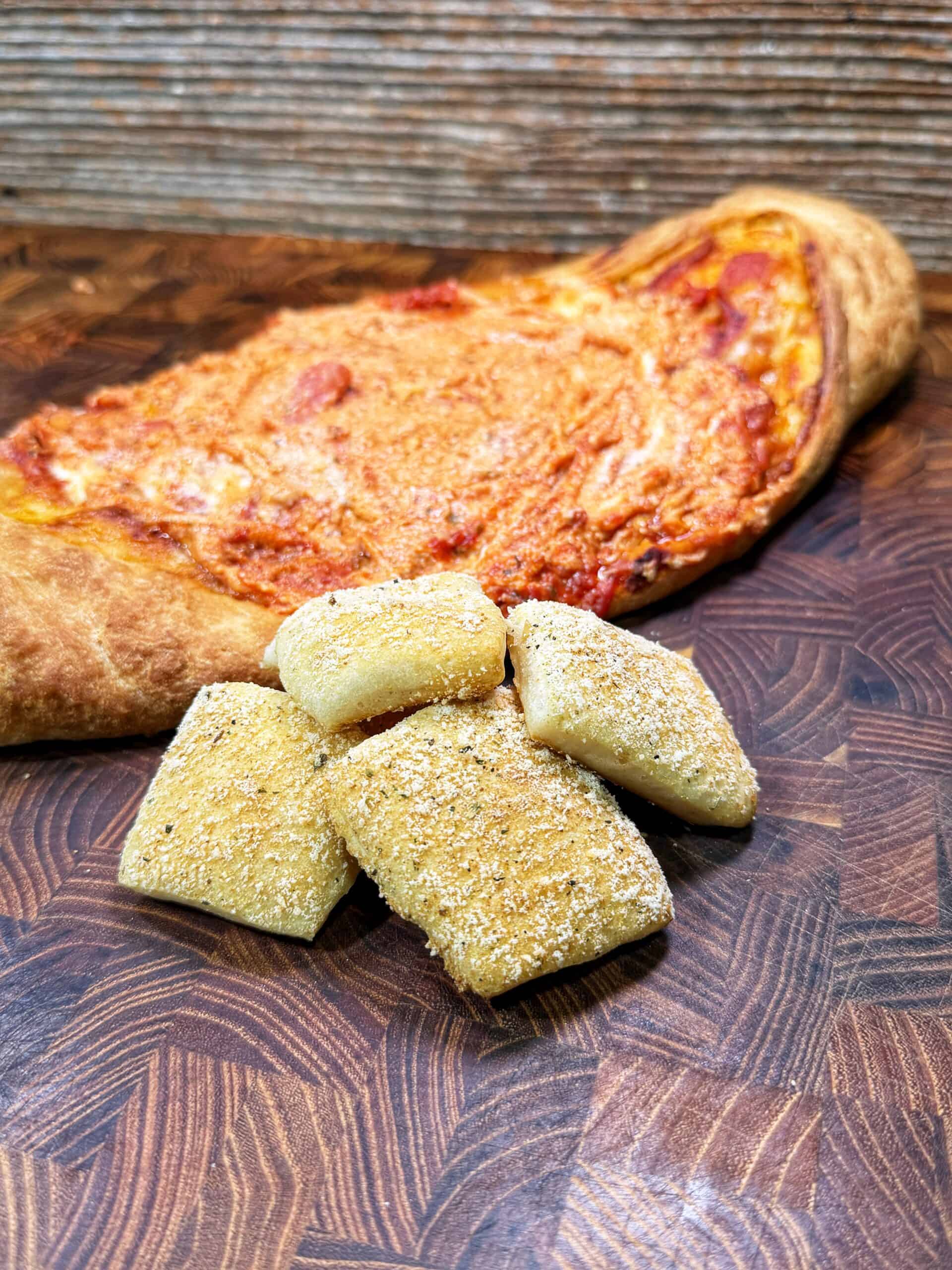 A golden brown calzone sits on a wooden cutting board, with several rectangular pieces of bread dusted with seasoning placed in front of it.