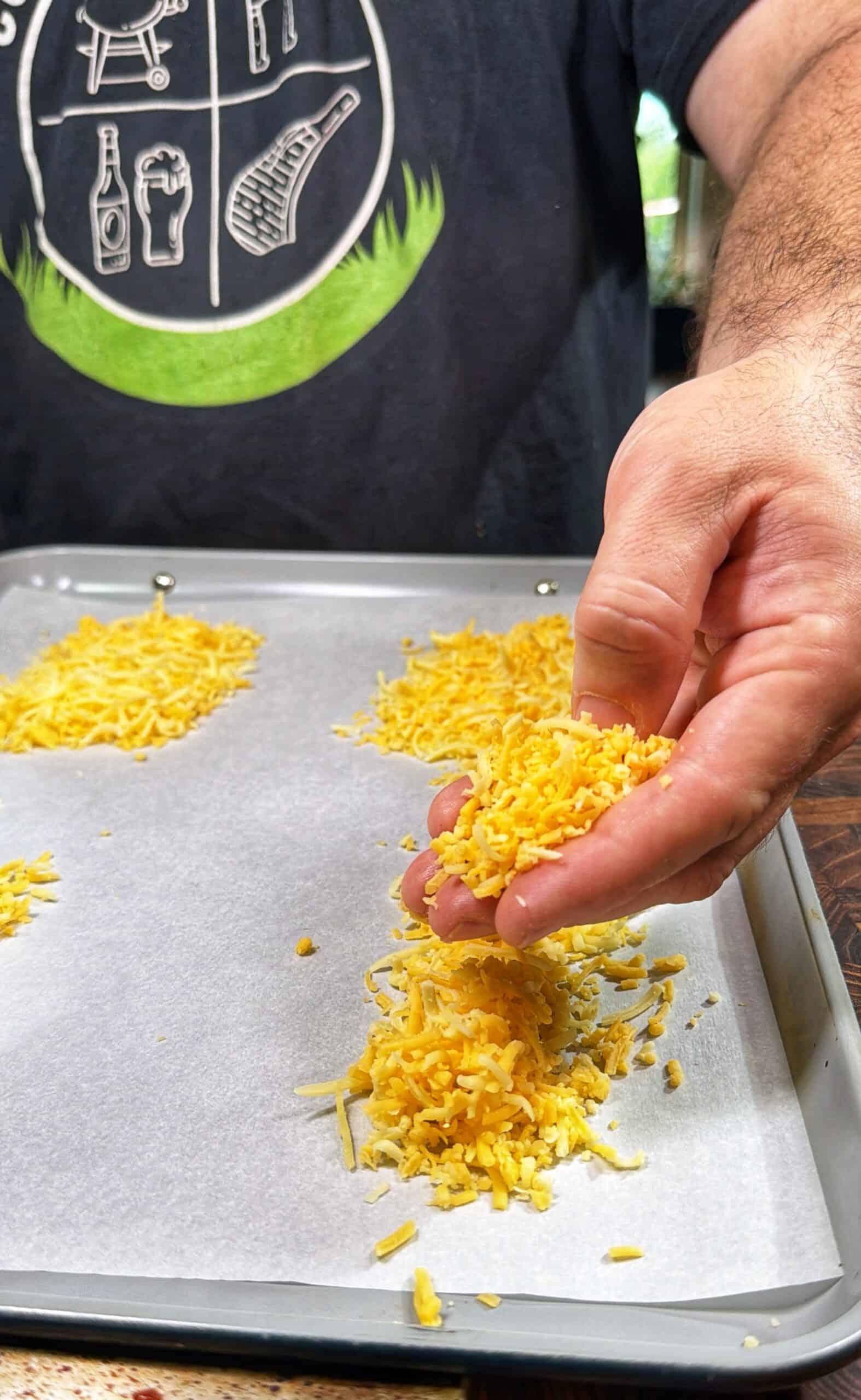 A person sprinkles shredded yellow cheese onto a parchment-lined baking sheet, preparing several small piles. The person is wearing a black shirt with graphic designs.