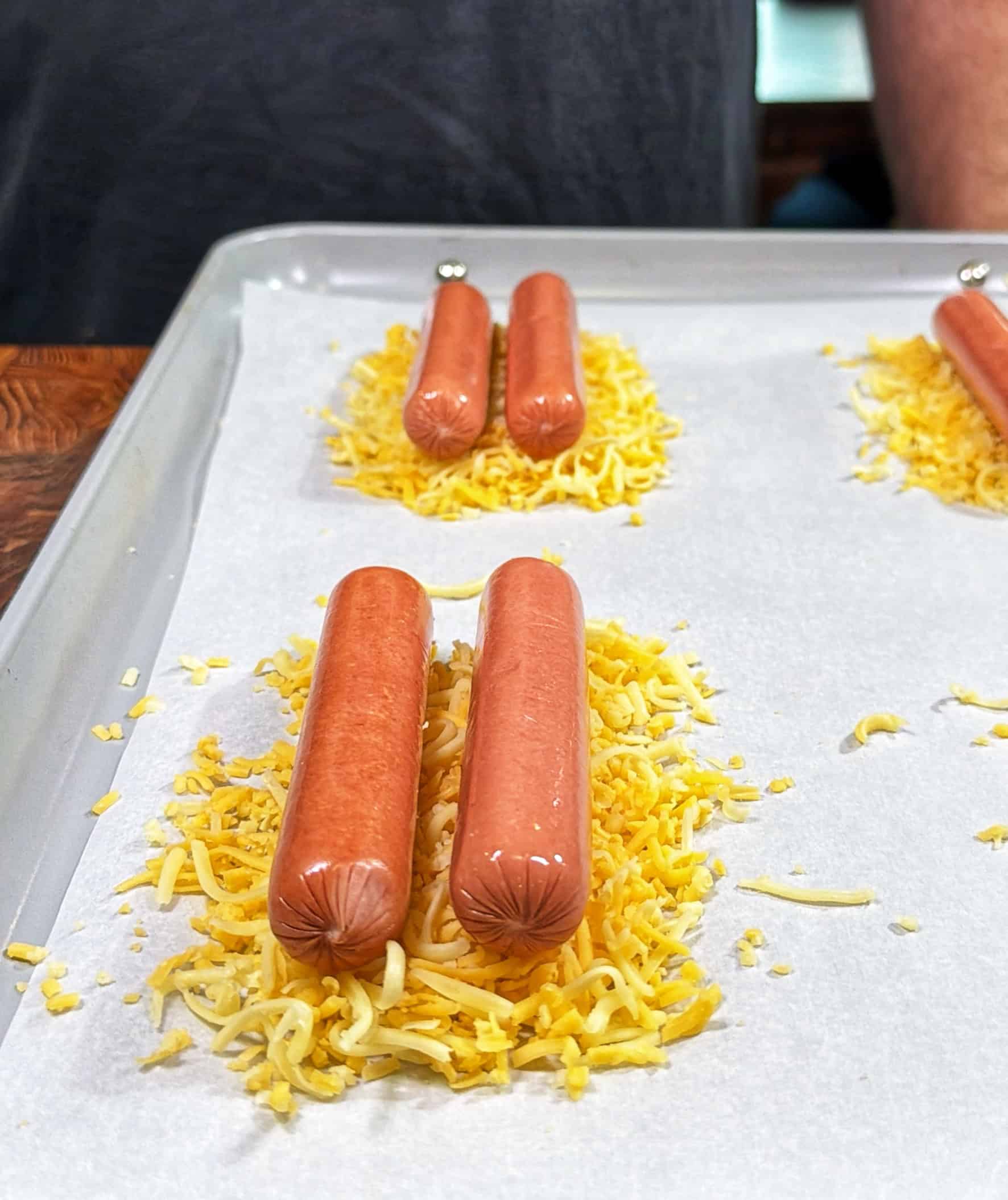 Two uncooked hot dogs are placed parallel on a small pile of shredded cheese on a parchment-lined baking sheet, with several similar arrangements in the background.