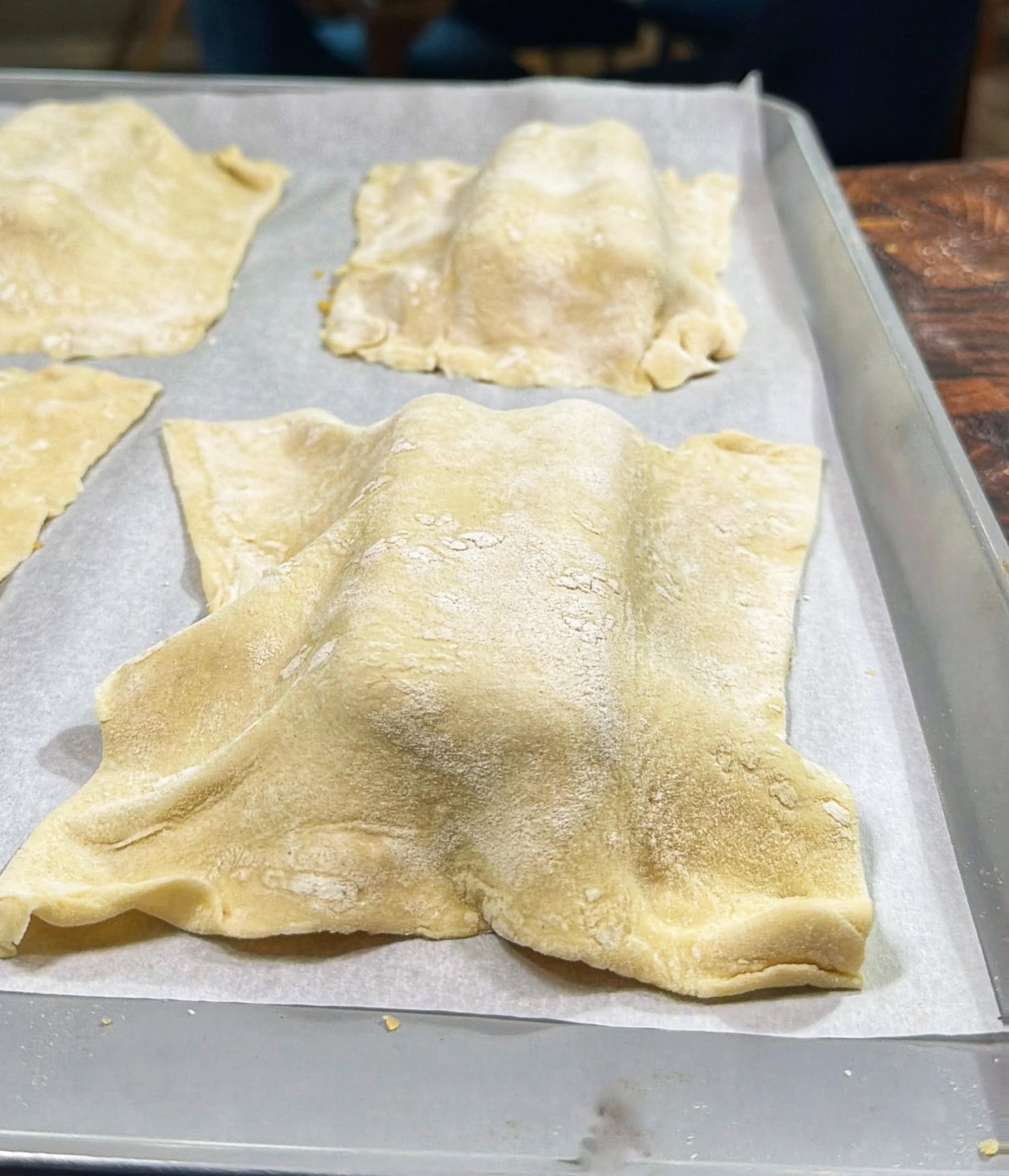 Sheets of fresh pasta dough covering mounds of filling rest on parchment paper atop a baking tray, ready to be shaped into ravioli or another stuffed pasta.