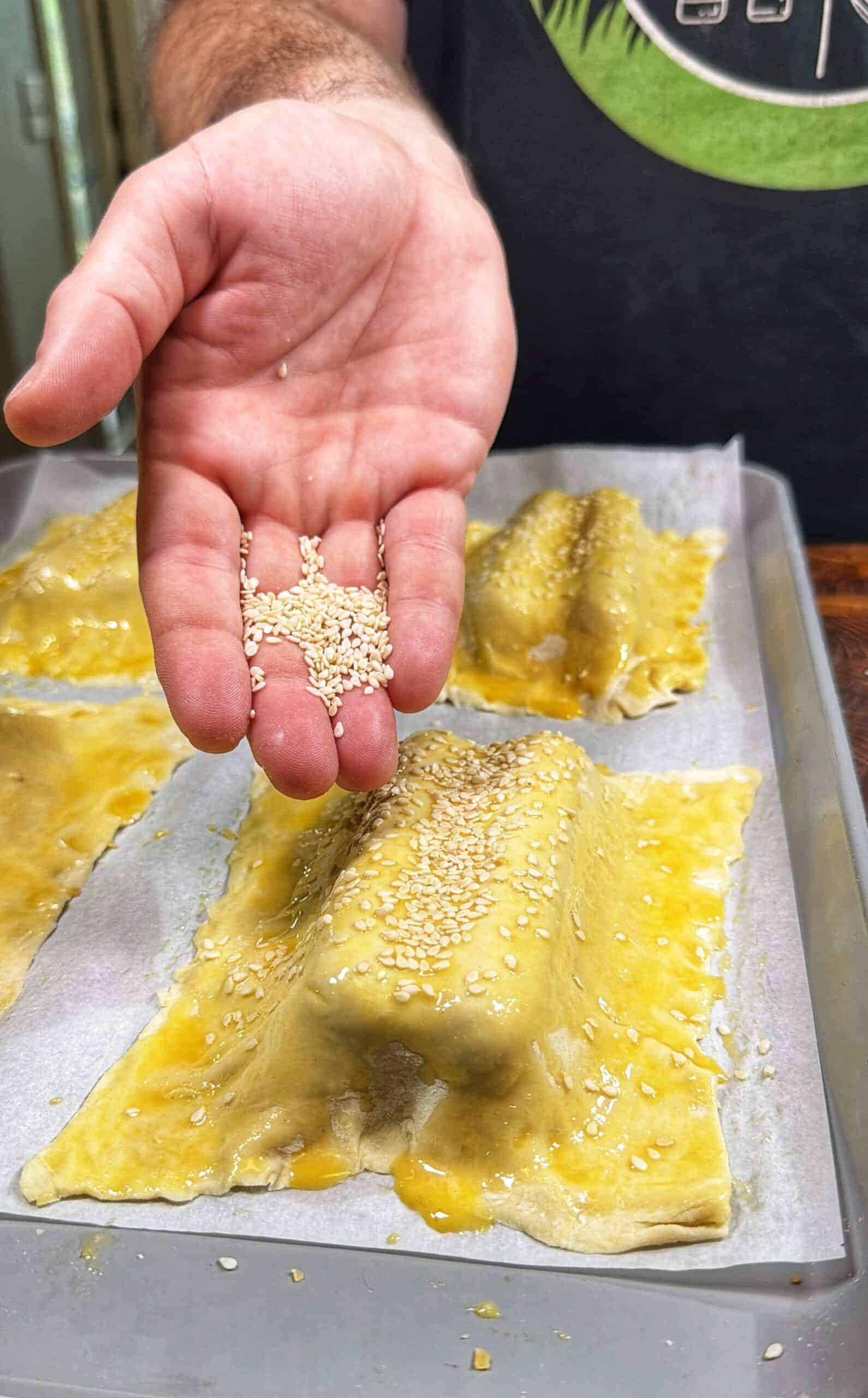 A hand sprinkles sesame seeds over rectangular, egg-washed pastry dough pieces on a baking tray lined with parchment paper.