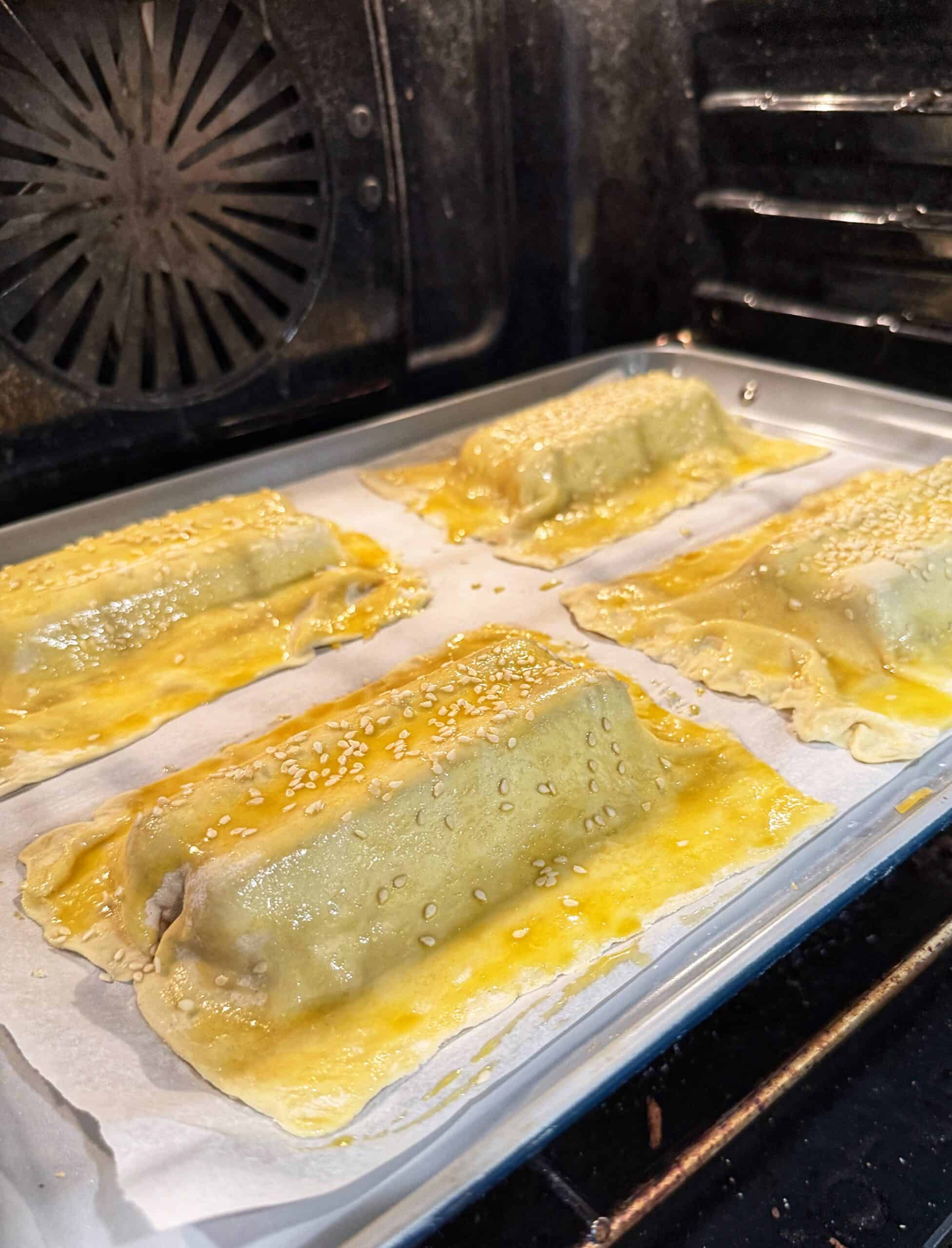 Rectangular pastry parcels topped with sesame seeds are baking on a parchment-lined tray inside an oven. The pastries are golden brown and appear to be brushed with egg wash.
