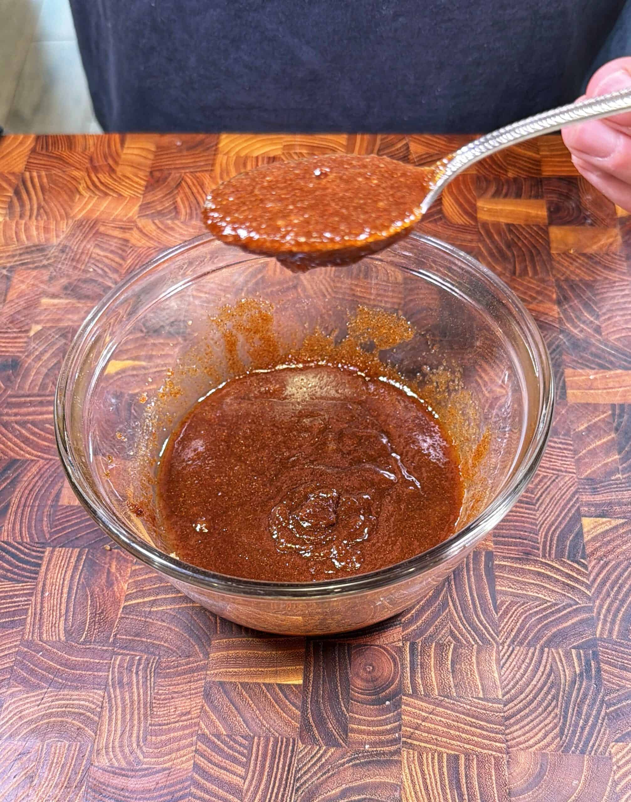 A person holds a spoon above a glass bowl filled with a reddish-brown spice mixture or sauce, set on a wooden countertop.