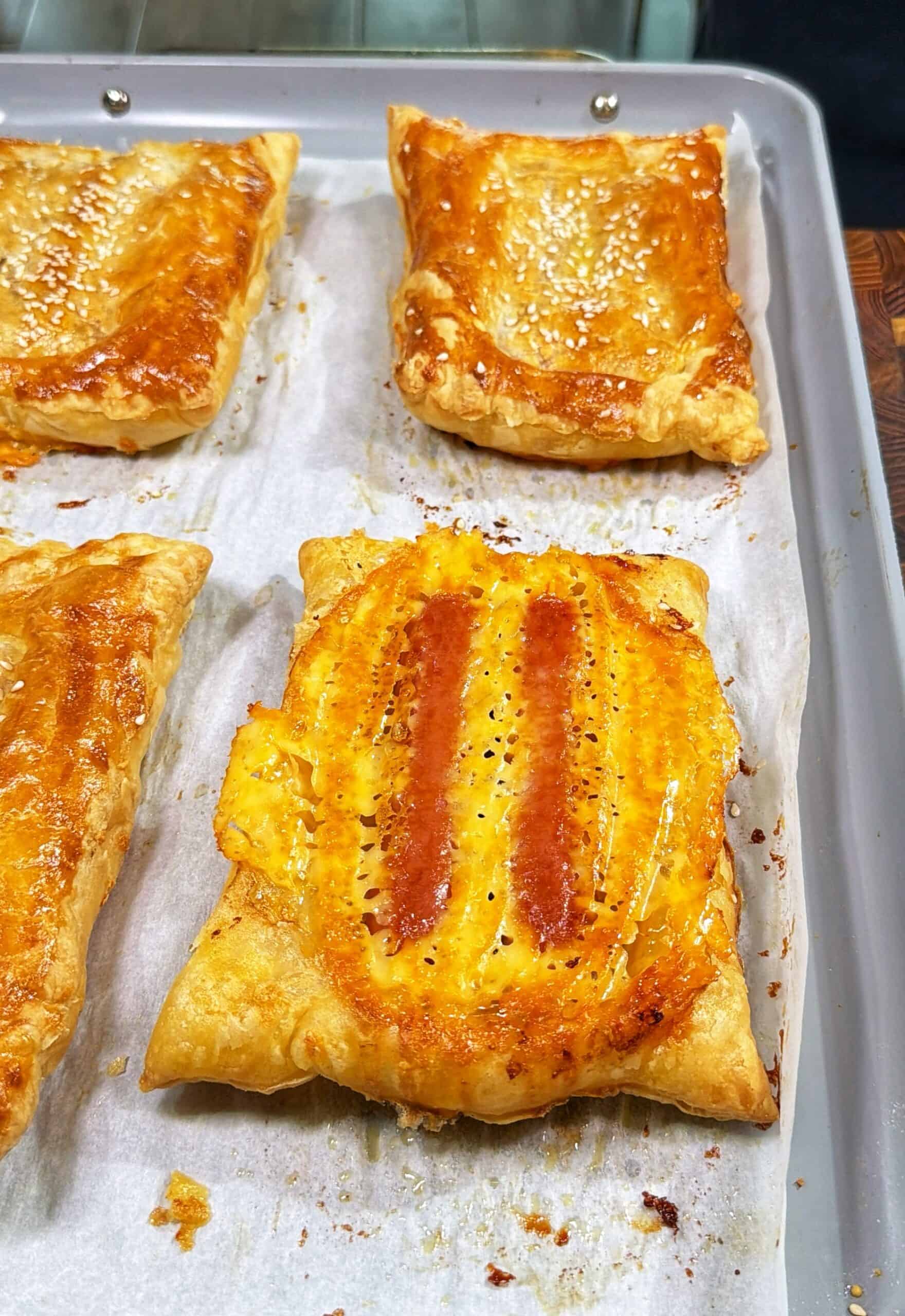 Four golden brown puff pastry squares on a parchment-lined baking tray; two have a shiny, egg-washed top with sesame seeds, and two display a more textured, oval marking in the center.