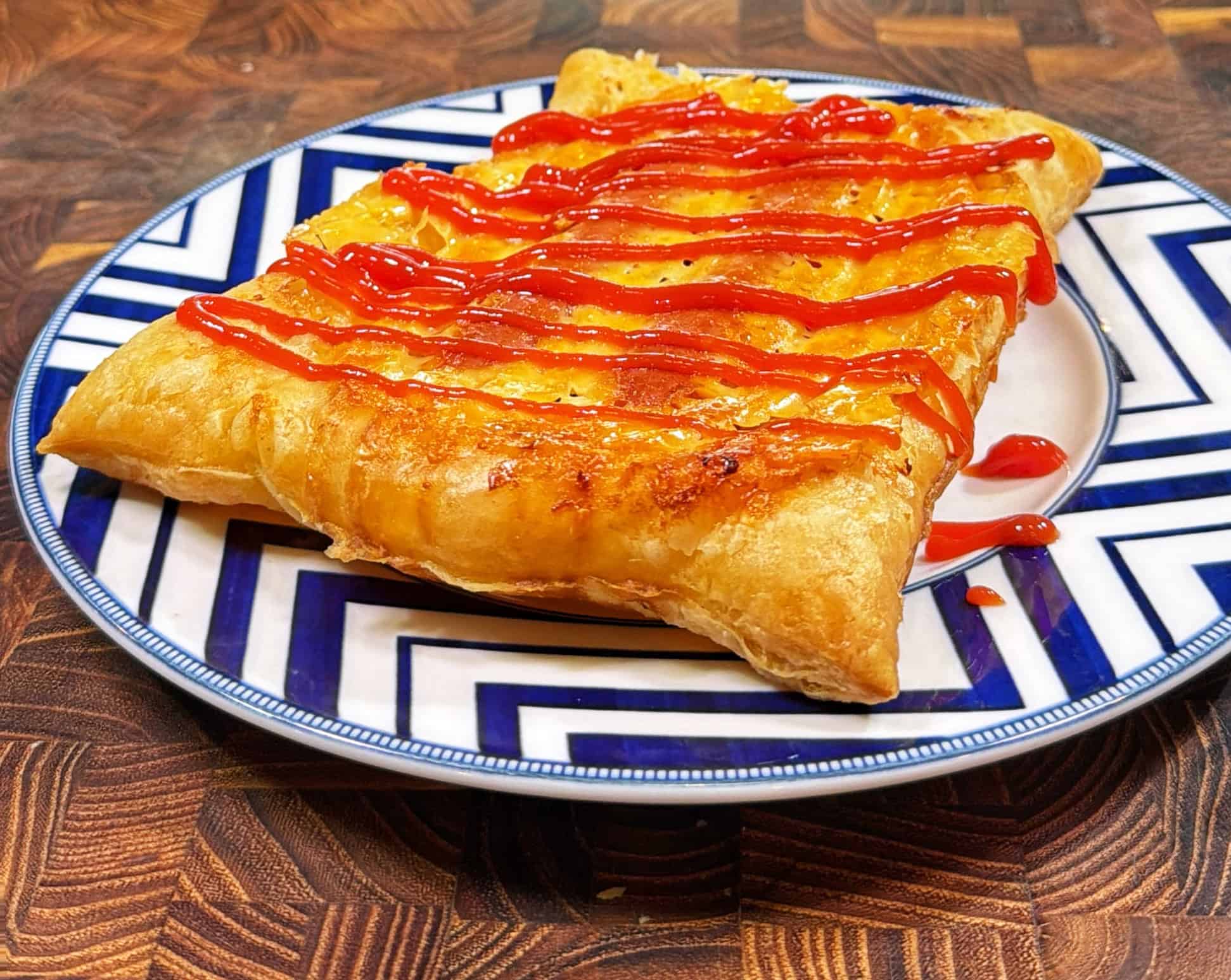 A flaky, golden-brown pastry topped with zigzag lines of ketchup sits on a decorative blue-and-white plate, placed on a wooden surface.