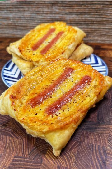 Two rectangular puff pastries with a golden, flaky crust and two reddish filling stripes on top are placed on a patterned plate and a wooden surface. A rustic wooden panel serves as the background.